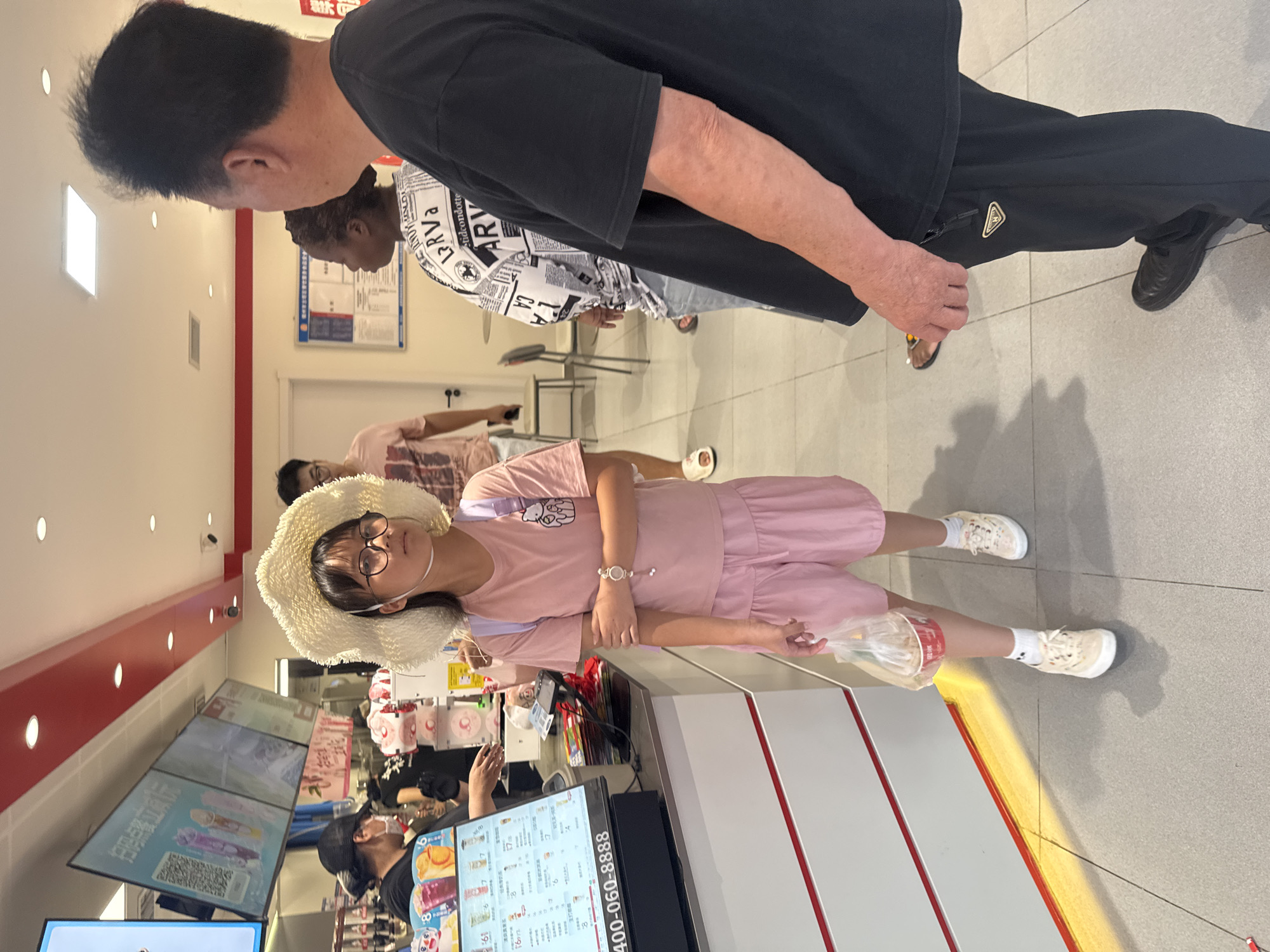 In Zhongtun, China, a young girl wearing a large straw hat and glasses stands in line at a food establishment. She is dressed in a pink t-shirt and matching shorts, with white sneakers and socks. She holds a clear plastic bag containing a small red cup. To her right, a man in a black t-shirt and black pants stands with his back to the camera. Further back, a woman with dark hair and a patterned shirt is visible. In the background, a digital menu displays various drinks. The environment appears to be indoors, with bright lighting and tiled flooring.