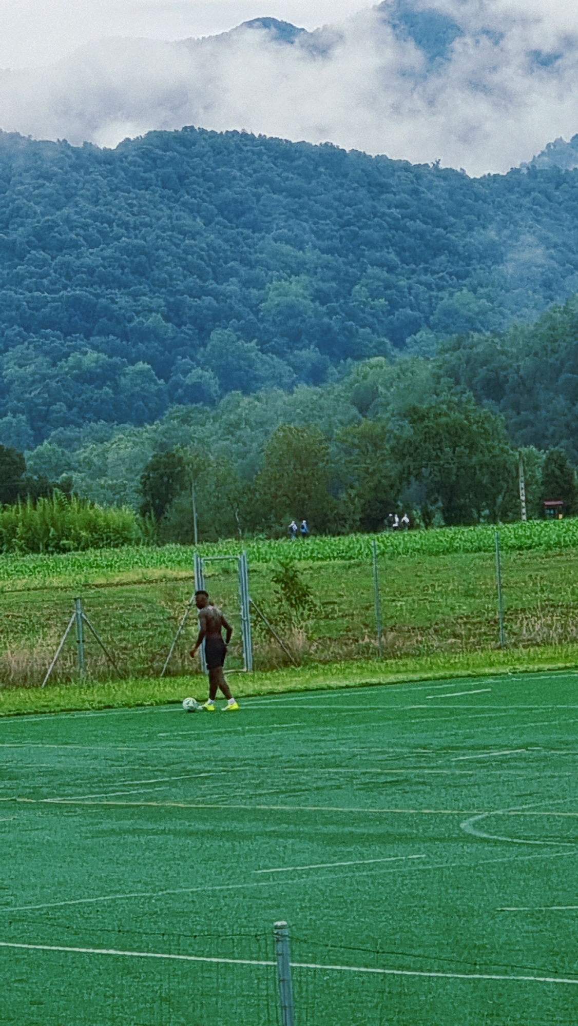 In La Canya, Spain, a young Black male, shirtless and wearing black shorts and bright yellow soccer cleats, stands on a green artificial turf soccer field. He is positioned to the left of the frame, facing slightly right, with a white soccer ball at his feet. In the background, a chain-link fence separates the field from a grassy area with some scattered trees and a distant field of crops. Beyond the field, a lush, densely forested mountainside rises, its slopes covered in a variety of green trees. The sky above is overcast with white clouds, obscuring the very top of the mountains. Several other individuals, appearing as small figures, are visible in the middle ground, near the fence line. The overall scene depicts an outdoor training session or casual game on a cloudy day.