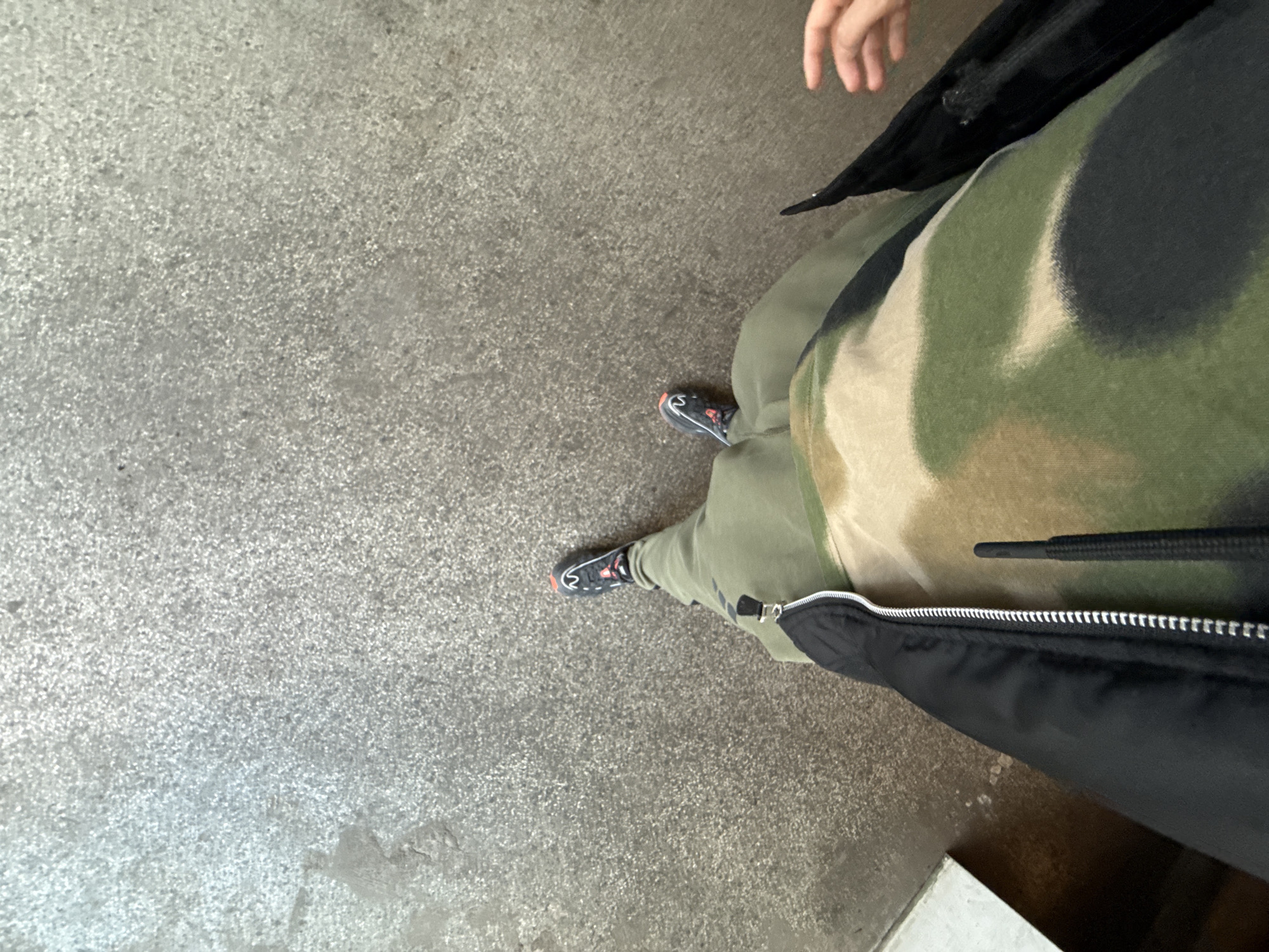 The image depicts a person standing on a gray concrete surface, likely a sidewalk or pavement. The person is wearing camouflage pants and black sneakers with red accents. A black jacket is partially visible, and the person's fingers are visible in the upper right corner. The background is a gray concrete surface. There is no indication of the image's location being Stara Zagora, Bulgaria.