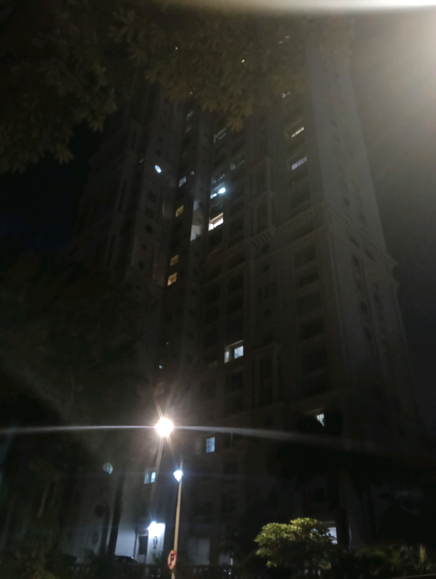 This is a nighttime, low-angle shot of a tall, illuminated building in Mumbai, India. The building is modern, with many windows, some of which are lit. In the foreground, there is a streetlamp and some foliage. The image appears to have been taken from a moving vehicle, as indicated by the light trails across the frame. The sky is dark and overcast, and the overall mood is somewhat eerie.