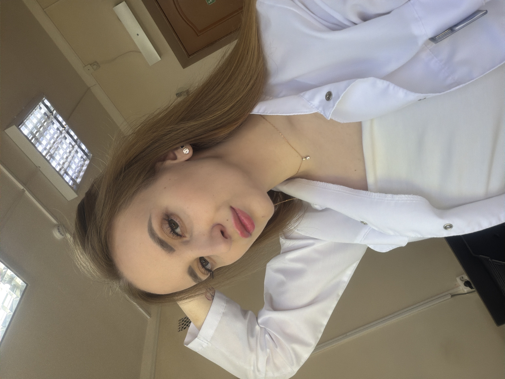 A young woman with light brown hair is posing for a selfie indoors. She is wearing a white lab coat over a white top and a delicate gold necklace. Her makeup includes defined eyebrows, mascara, and pink lipstick. She is looking at the camera with a slight smile.
The background includes a ceiling with fluorescent lights, a wooden door, and beige walls. Based on visual cues such as the interior design and the presence of a lab coat, the location is likely a medical or educational facility in an urban area. There is no definitive evidence in the image to place it specifically in the city of Chaikovsky, Russia.