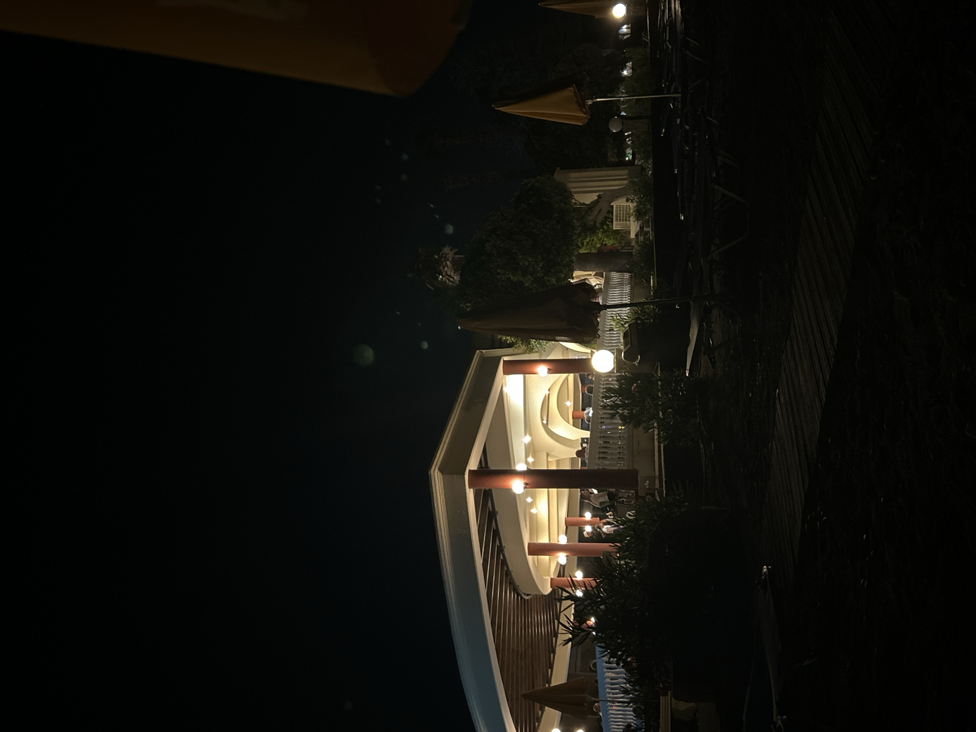 This is a nighttime, low-angle, full-shot photograph of a resort's outdoor dining area and beach chairs on the island of Rhodes, Greece. In the background, a curved, open-air pavilion with a slatted wooden roof is illuminated by numerous warm, spherical lights. The pavilion has white balustrades and is supported by reddish-orange columns. People are visible inside the pavilion, appearing to be dining or socializing. In the foreground, dark silhouettes of lounge chairs and beach umbrellas are arranged on the sand, with a wooden walkway in the very front. Several small, round lights are visible near the ground in front of the pavilion. The sky is dark, with a few indistinct blurry lights, possibly lens flares or atmospheric phenomena. The overall impression is of a tranquil, after-hours resort ambiance.