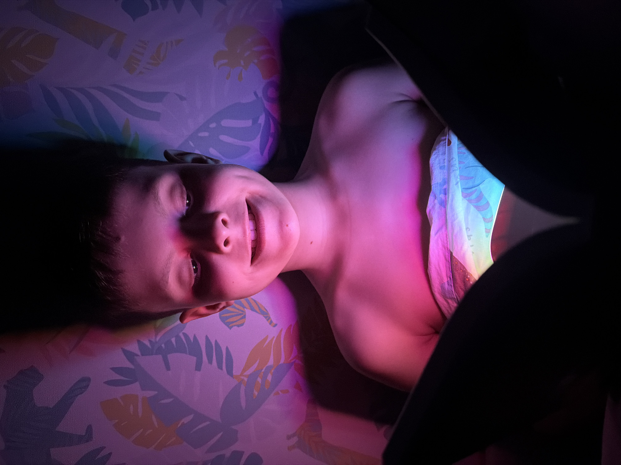 A person lies on their back on a patterned surface, possibly a bed, illuminated by a mix of pink, purple, and blue light. The person is smiling slightly and looking toward the camera. A black object, partially obscuring the view, is positioned on the right side of the frame. The surrounding environment is dark. The setting appears to be indoors, and based on the supplied crops there is no verifiable information to support the image being taken in Votkinsk, Russia.