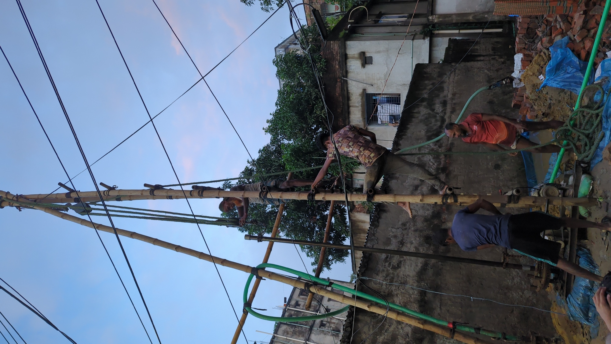 In Dhaka, Bangladesh, four men are engaged in a construction activity involving a bamboo scaffolding structure and a drilling rig. The scaffolding, made of bamboo poles lashed together with ropes, supports a pulley system. Two men are positioned on the scaffolding, one above the other, appearing to operate the machinery. The lower man on the scaffolding is wearing a patterned shirt and dark pants, while the man above him is wearing a light-colored tank top and dark shorts.
Below the scaffolding, two other men are working. One man, wearing a blue t-shirt and black shorts, stands by the drilling rig, which is partially visible at the bottom center of the image. He is looking down at the machinery. Another man, wearing a red t-shirt and checkered shorts, stands to the right of the drilling rig, observing the operation. He is holding a green hose.
The environment appears to be an urban construction site. A partially constructed building with a grey concrete wall is visible in the background, along with some trees. Electrical wires crisscross the sky, which is a pale blue with some hints of pink, suggesting either early morning or late afternoon. The ground is covered with dirt, mud, and piles of bricks. A blue tarp is spread out on the ground, partially covered with soil and debris. The overall scene depicts manual labor and construction work in a developing urban area.
