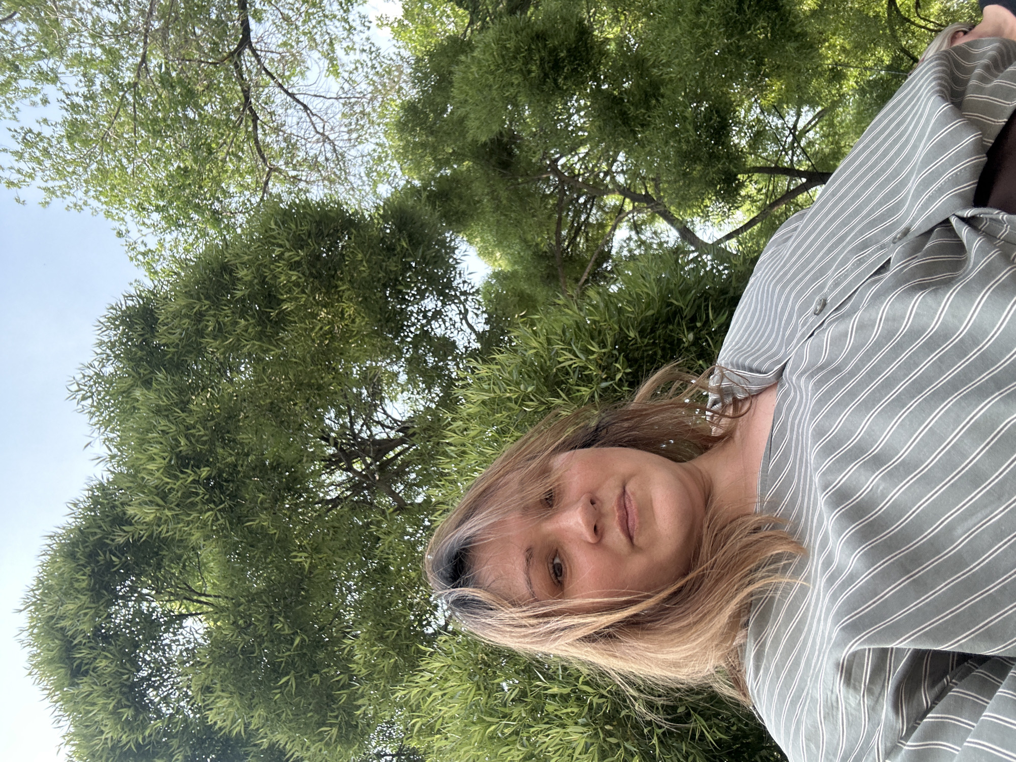 In Omsk, Russia, a woman with light brown hair and bangs is captured in a low-angle, outdoor selfie. She is looking towards the camera with a neutral expression, and her face is partially obscured by her hair. She is wearing a light green, striped shirt. The background is dominated by lush green trees and a clear sky, suggesting a natural, possibly park-like setting within the city. The perspective of the shot is from below, looking up at the subject and the surrounding foliage.