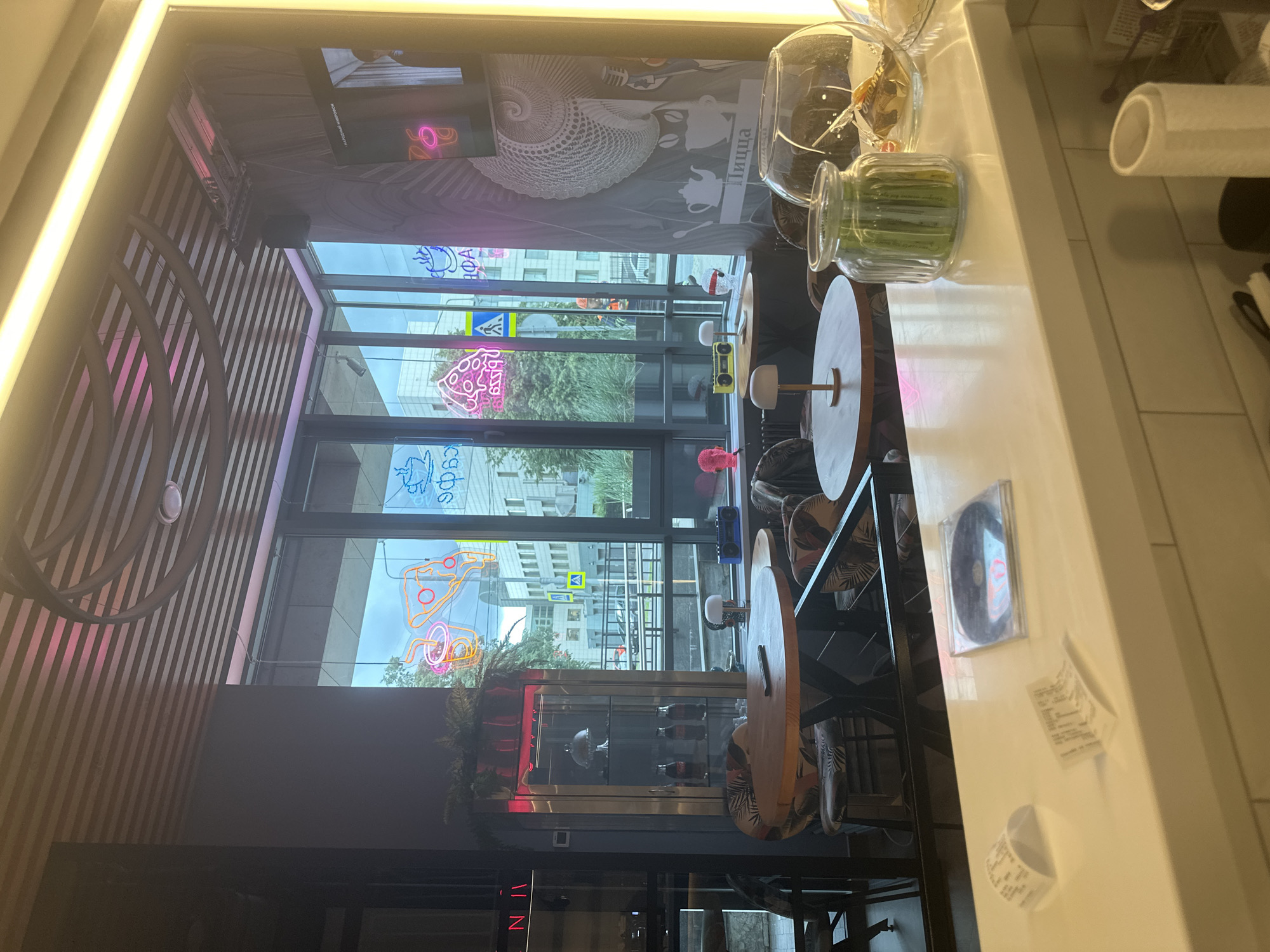 The image depicts the interior of a cafe, likely located in Перовское сельское поселение, Ukraine. The scene is brightly lit, with a prominent neon sign visible through the large windows, which offer a view of the city street outside. Inside, several round tables with chairs are arranged, some of which are occupied by customers. A counter area is visible in the foreground, with various items placed on its surface, including receipts and a CD case. The cafe's decor appears modern and vibrant, with artistic wall decorations and a unique ceiling design. The overall atmosphere suggests a bustling and contemporary establishment.