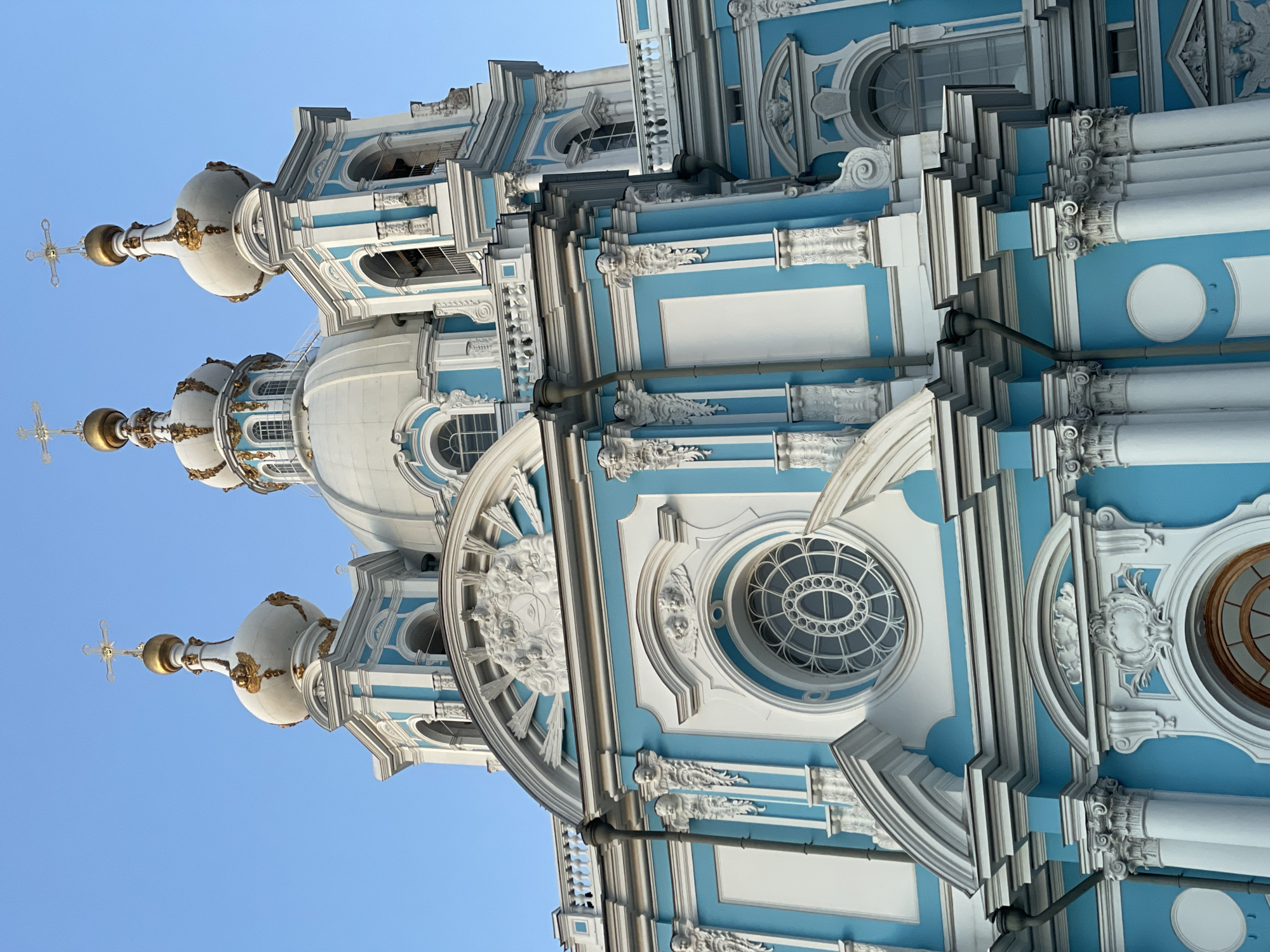 This is a low-angle, close-up, eye-level, outdoor, medium shot of the Smolny Cathedral in Saint Petersburg, Russia. The sky is clear and blue. The cathedral is ornate, with white and light blue stucco walls and golden domes. The architecture is Baroque, characterized by elaborate ornamentation, including white stucco reliefs of cherubs, floral motifs, and a large circular window with intricate tracery. Multiple domes and cupolas, topped with crosses, rise above the structure. The building features white columns and decorative moldings. The scene is static, with no people or animals visible.