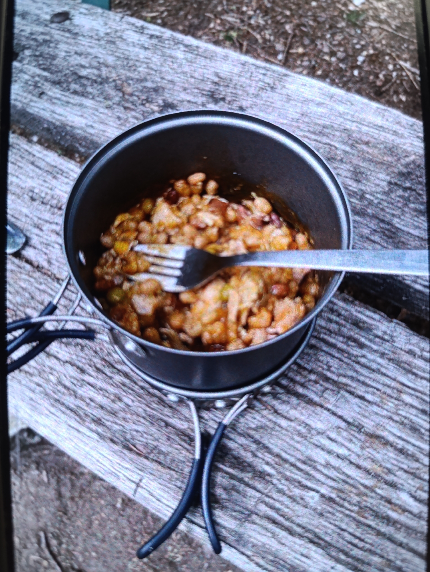 A close-up, slightly out-of-focus overhead shot shows a camping pot filled with food resting on a weathered wooden picnic table. A metal fork is partially submerged in the food. The pot has two black handles that attach to a silver base. The picnic table is outdoors and appears to be in a natural setting, possibly a park or campsite, with some dirt and debris visible in the background. The lighting suggests it is daytime.
