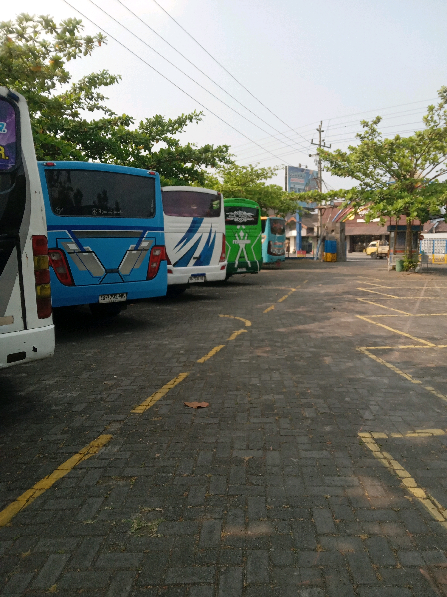 A row of parked buses is visible on a paved lot in Banguntapan, Indonesia. The buses are various colors, including white, blue, and green, and are parked in designated spots marked by yellow lines. Trees with green foliage line the background, and a clear sky is overhead. A power line runs across the sky. In the distance, there are buildings and a small yellow truck. The ground is made of dark gray bricks.
