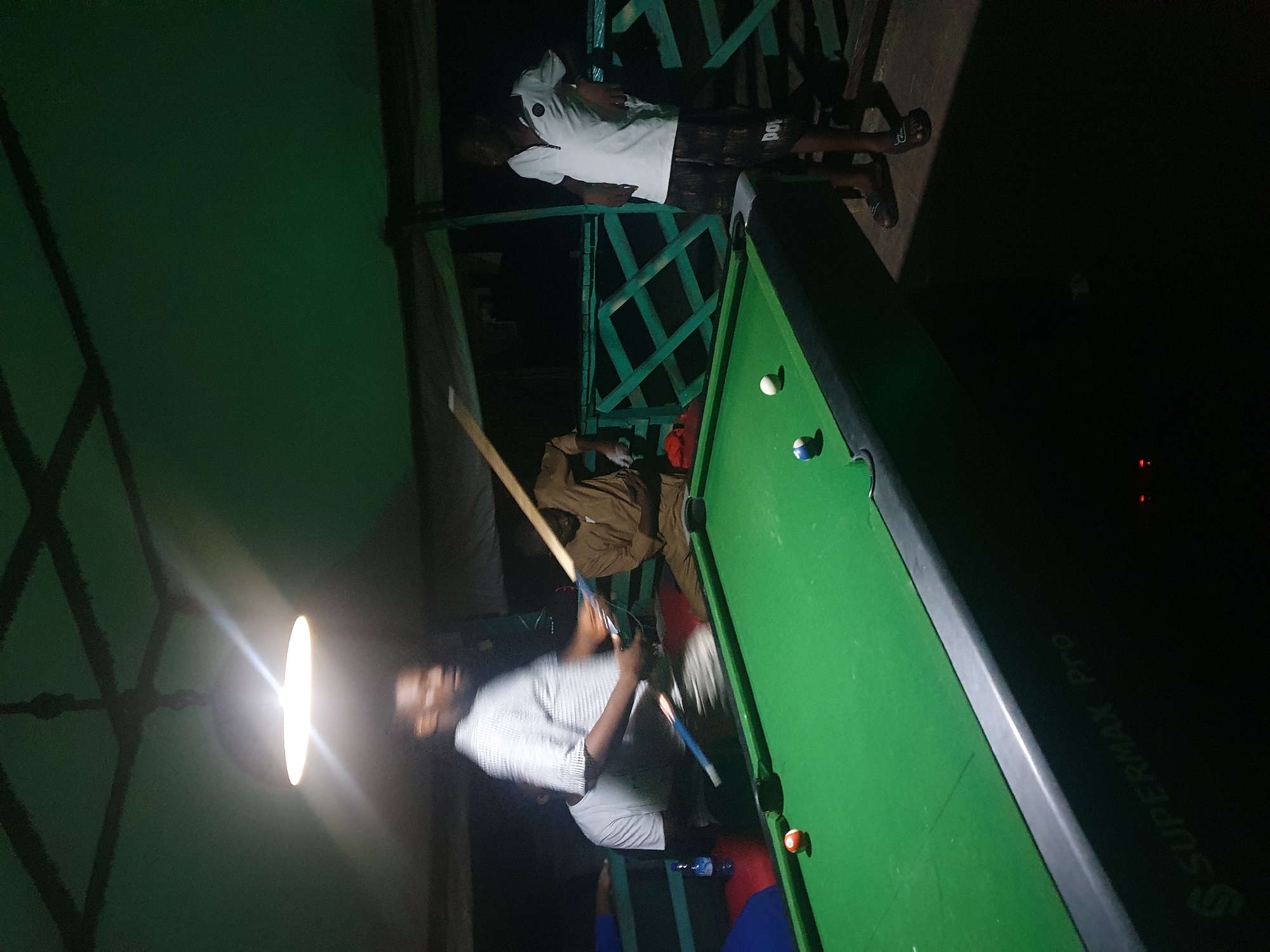 The image depicts an indoor billiard game taking place in Lokoja, Nigeria. Four men are present, with two actively playing. One man, wearing a checkered shirt, is positioned behind the billiard table, holding a cue stick and appearing to take a shot. Another man, dressed in a light brown traditional garment, is seated near the table, observing the game. A third man, in a white polo shirt and patterned shorts, stands to the right, leaning against a railing. The fourth man, partially visible in a white t-shirt, is on the left side of the table. The billiard table is green, with several balls visible on its surface. The setting appears to be a covered outdoor area, possibly a patio or veranda, with a ceiling fan and a light fixture illuminating the scene. The background shows a green wooden railing. The overall atmosphere suggests a casual gathering for a game of billiards.
