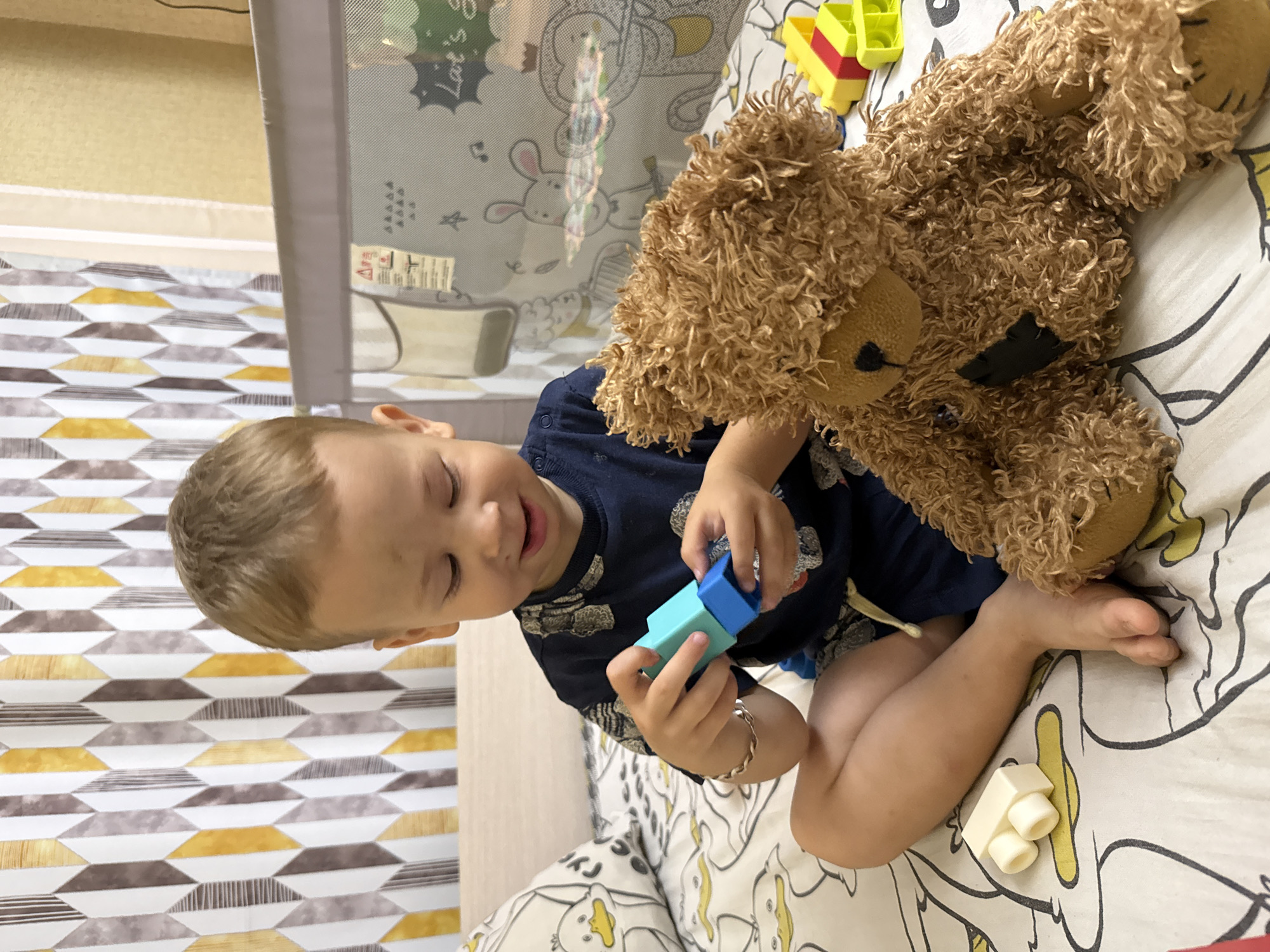 In Rusanovo, Russia, a young boy, estimated to be around 2-3 years old, sits on a bed with a patterned duvet featuring duck illustrations. He is wearing a dark blue t-shirt with a repeating pattern and blue shorts. The boy is engaged in playing with building blocks, holding a blue and a turquoise block in his hands. To his right, a large, shaggy brown teddy bear is positioned on the bed. In the background, a crib with a mesh siding is visible, decorated with images of animals and text. A patterned curtain with yellow, brown, and white geometric shapes hangs on the wall behind the crib. The overall setting appears to be a bedroom.