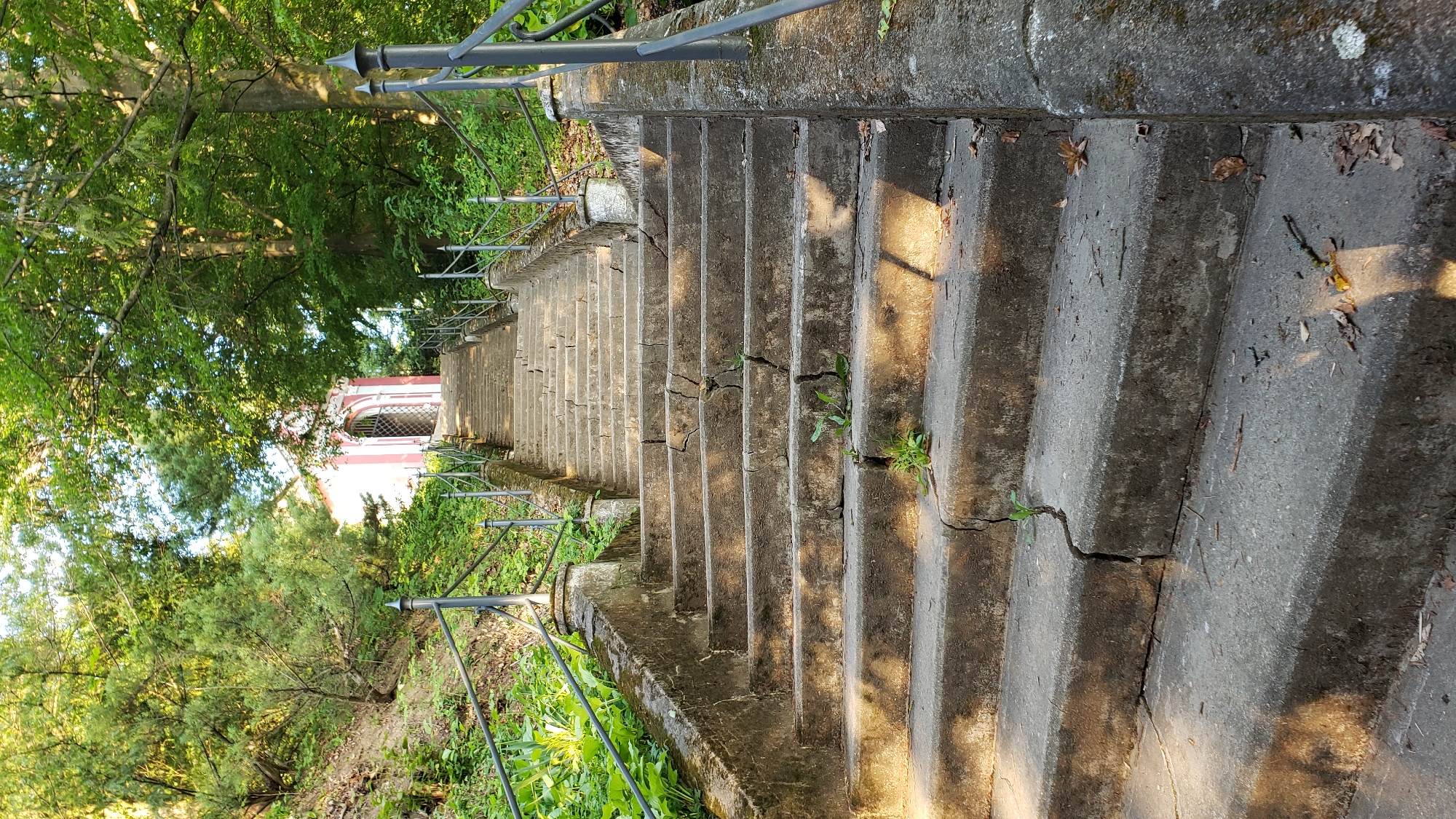The image depicts an outdoor scene featuring a long, worn concrete staircase leading upwards. The staircase is flanked by metal railings supported by stone pillars. Lush green foliage and trees surround the stairs, with sunlight dappling the stone steps. At the top of the stairs, a small, light-pink building with a decorative arched doorway is visible, suggesting it could be a chapel or a small monument. The overall impression is one of a serene, historical pathway in a natural setting. This staircase is located on the Kalvária hill in Prešov, Slovakia, a popular religious and recreational site. There are no people visible in the image.