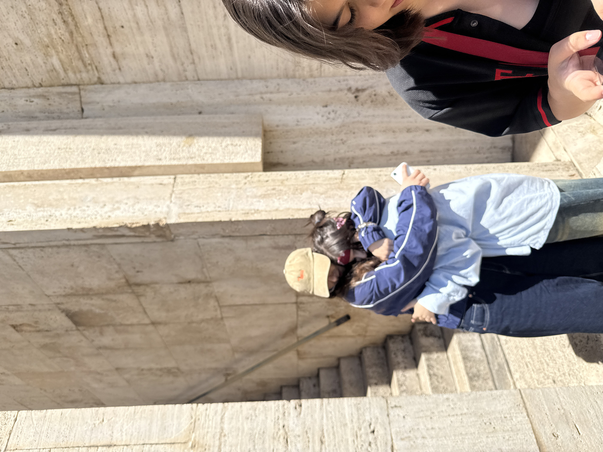 The image depicts a scene in Yerevan, Armenia. Two people, one wearing a tan baseball cap and a dark blue jacket, are standing on a light-colored stone staircase. The other person wears a light blue shirt and dark blue jeans. They appear to be holding a white phone. Above them and slightly out of focus, a third person is visible with dark hair and a black shirt with red accents. The overall environment is dominated by the staircase and light stone architecture.