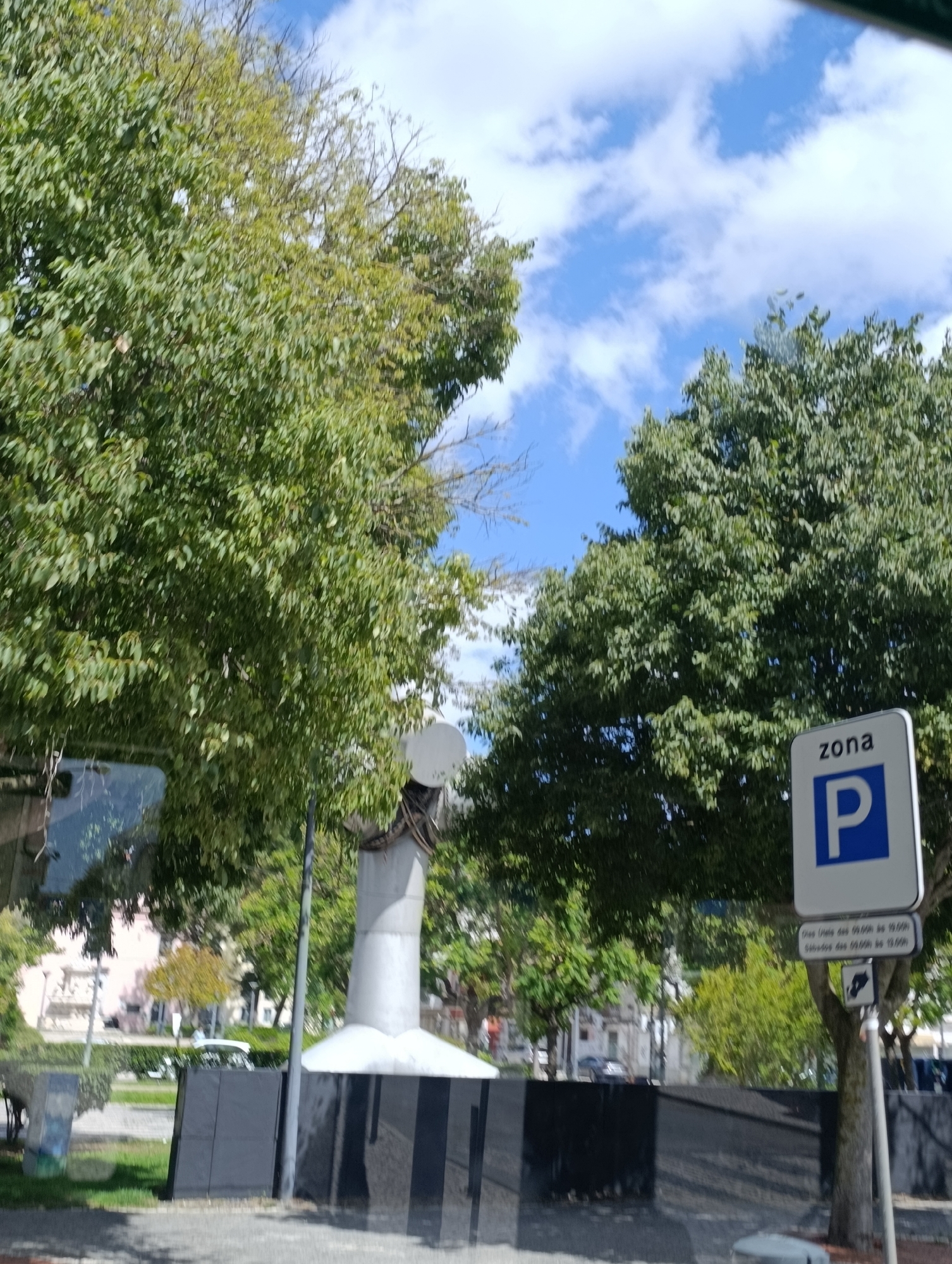 The image captures a street scene in Setúbal, Portugal, under a bright blue sky with scattered white clouds. Lush green trees dominate the foreground and midground, with their leaves creating a dense canopy. In the right midground, a white sign with a blue "P" inside a blue square is clearly visible, indicating a parking zone. Below this, another smaller sign provides information in Portuguese about the parking hours: "Dias Úteis das 09:00h às 19:00h" (Weekdays from 09:00 to 19:00) and "Sábados das 09:00h às 19:00h" (Saturdays from 09:00 to 19:00). A grey pole supports these signs. To the left of the parking signs, a tall, grey, cylindrical structure with a white conical top rises from a black, reflective base. This structure appears to be a sculpture or monument. Behind the trees and monument, buildings are partially visible, suggesting an urban environment. The foreground shows a paved area and some greenery. The overall impression is a sunny day in a city park or street with significant tree cover and public amenities.