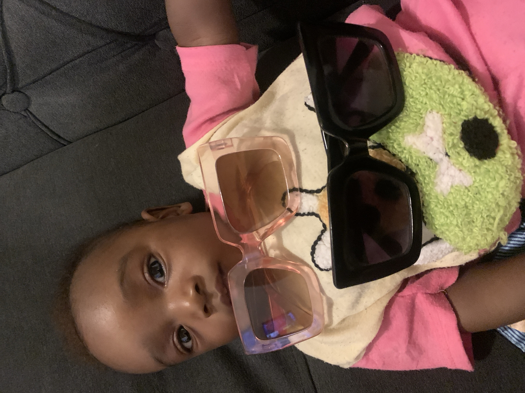 A young child lies on a dark grey, textured surface. The child is wearing a pink long-sleeved shirt over a yellow and white printed shirt with a green plush item with a white cross and black dot on it. Two pairs of sunglasses are positioned on the child's chest. One pair has a light pink frame, and the other has a black frame. The child is facing the camera with their head tilted, and their eyes are open. There is no evidence to suggest the location is Barakin Akawo, Nigeria.