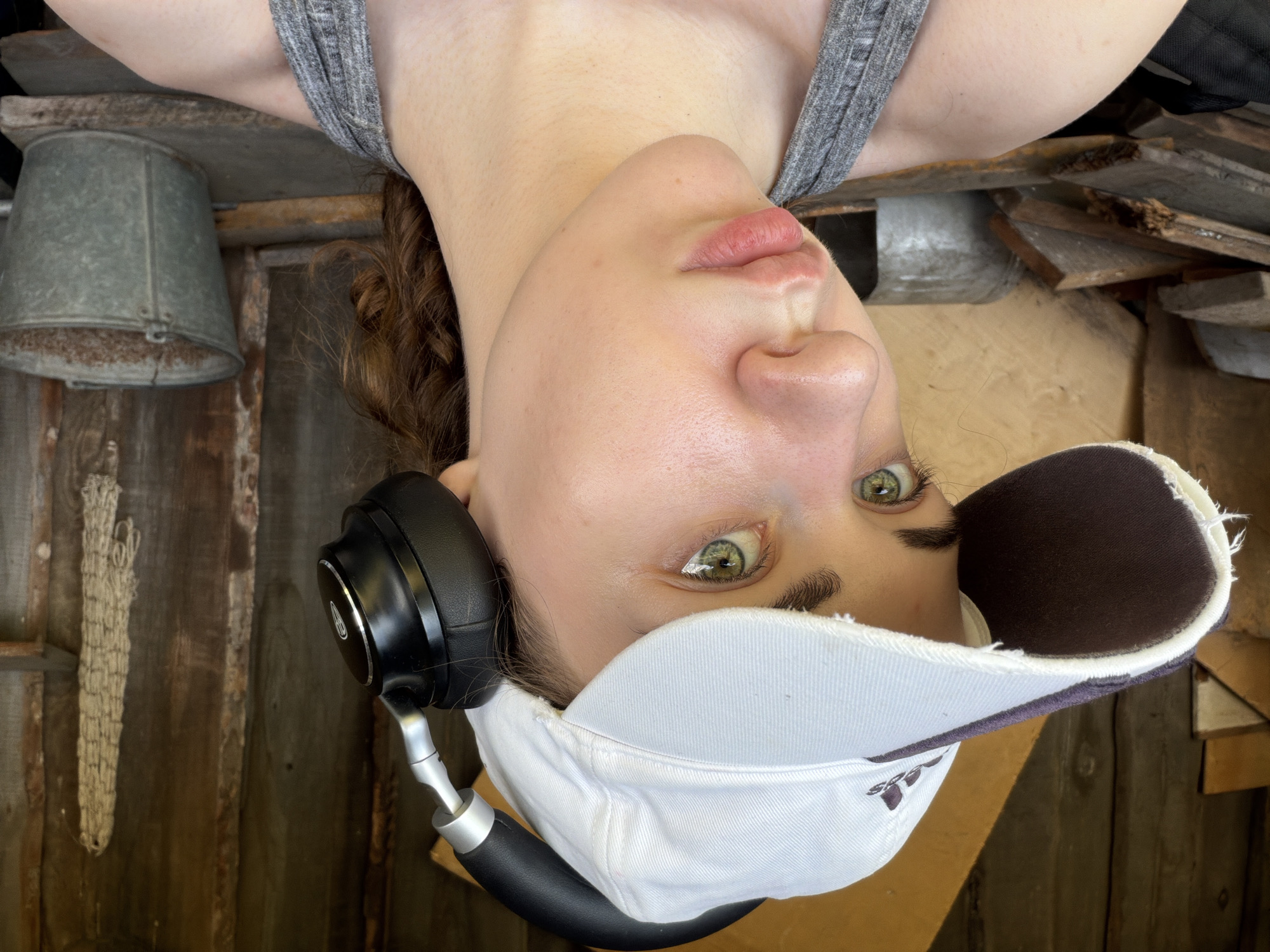 A young Caucasian female with light skin, green eyes, and reddish-brown curly hair pulled back in a ponytail is shown from the chest up, facing slightly to her left. She is wearing a white baseball cap with a dark brim and a grey tank top. Black over-ear headphones are resting on top of her cap. Her expression is neutral, with her lips slightly pursed. The background appears to be an outdoor, rustic setting, possibly a shed or outbuilding with wooden walls. Piles of lumber and a metal bucket are visible behind her. The scene is illuminated by natural sunlight, casting shadows. The image was captured in Zavodoukovsk, Russia.
