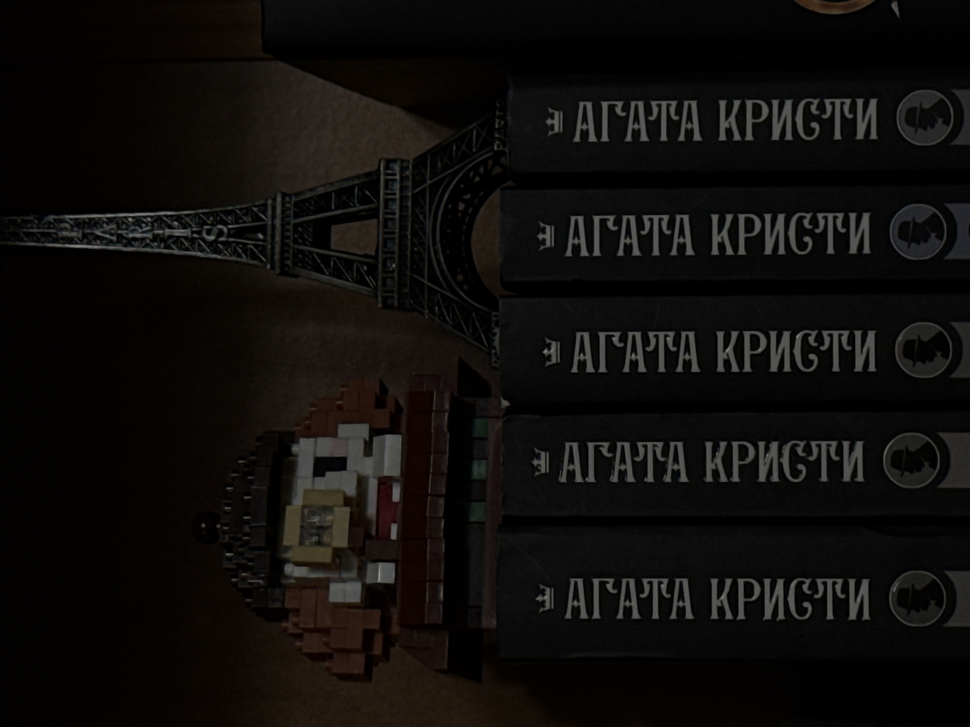 The image depicts a shelf with several items. On the left, there is a pixelated figurine of a person wearing a hat and a coat, with visible facial features. To its right, a miniature Eiffel Tower replica stands tall. Below these items, five black book spines are visible, each featuring white text that appears to be Cyrillic and a circular emblem with a silhouette of a person wearing a hat. The lighting is dim, creating shadows and highlighting the textures of the objects. The background appears to be a dark, textured surface, possibly a wall or another shelf. There is no indication of the city of Luhansk, Ukraine, or any specific location.