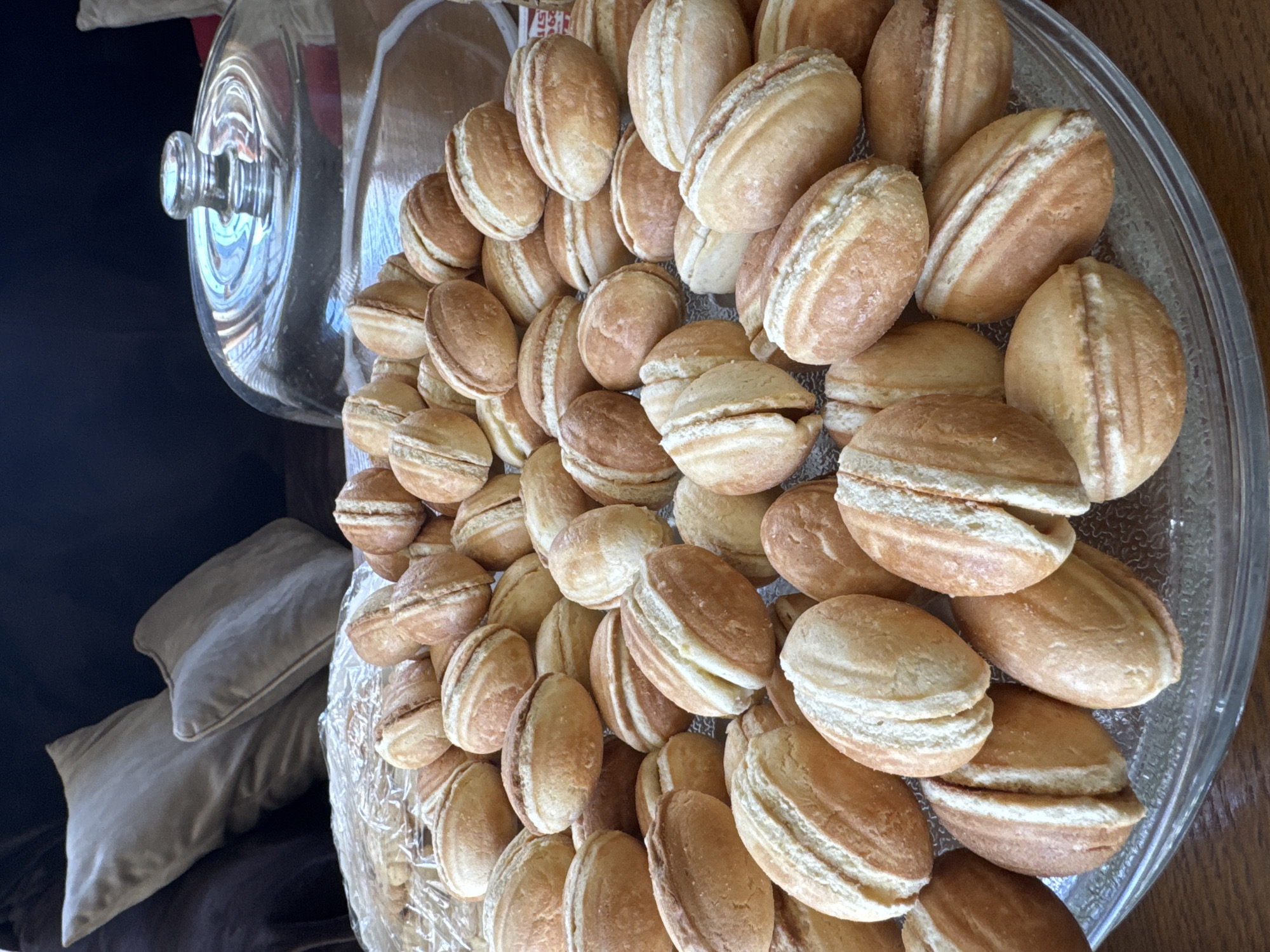 A glass platter filled with nut-shaped sandwich cookies sits on a wooden surface. The cookies are golden brown with a light filling. The platter has a domed glass lid, partially visible in the upper left corner. A dark blue cushion and a light brown pillow are in the background. The location is Burgas, Bulgaria.