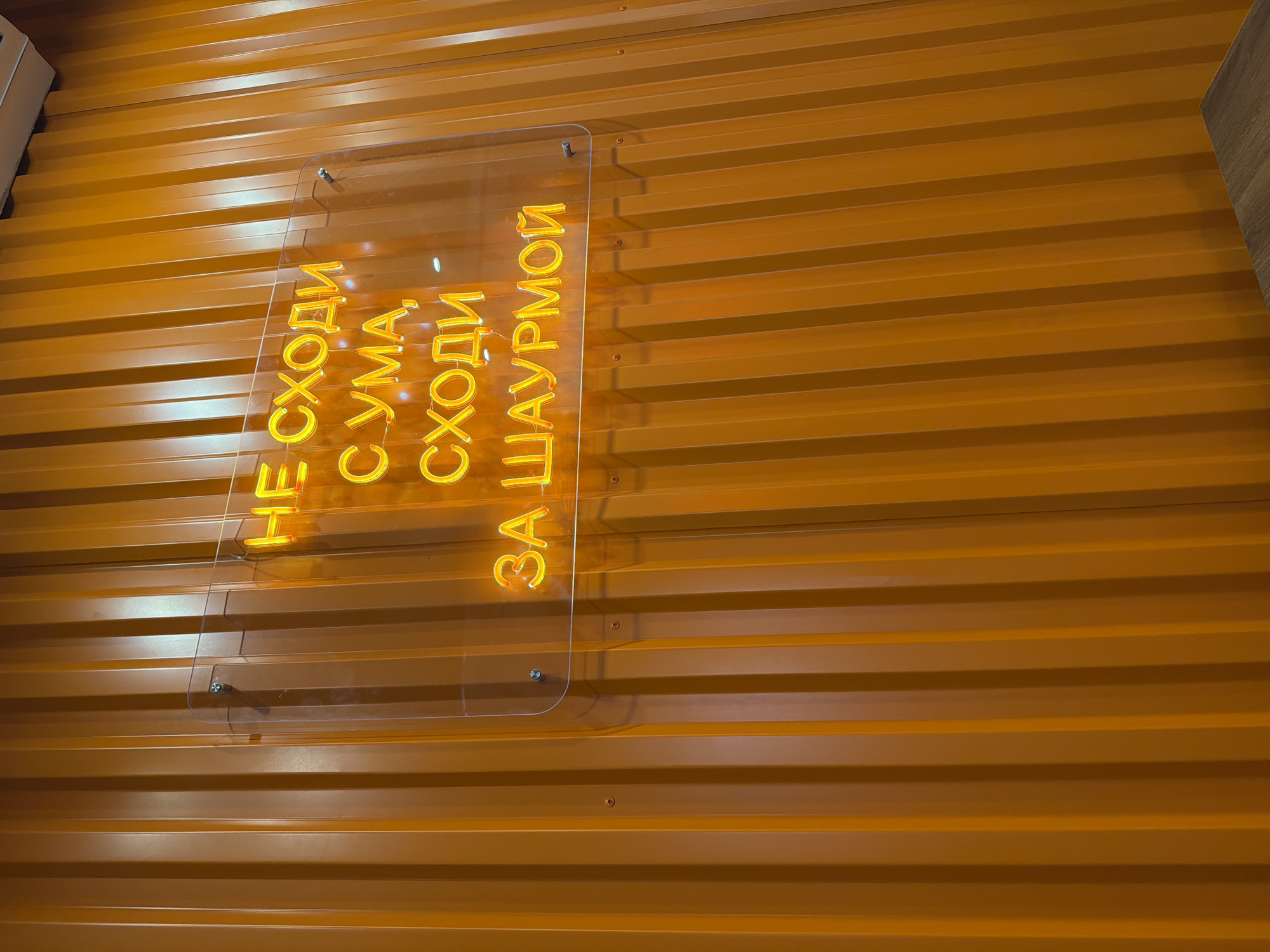 The image shows a close-up of an orange corrugated metal wall with a clear plastic sign attached to it. The sign features vertically arranged, illuminated yellow lettering in Cyrillic. The text reads "НЕ СХОДИ С УМА, СХОДИ ЗА ШАУРМОЙ." The sign is secured to the wall with clear mounting hardware. The wall is a uniform orange color with horizontal ridges. This appears to be an advertisement for Shawarma, though the location in the city of Krasnoyarsk is not explicitly stated in the image.