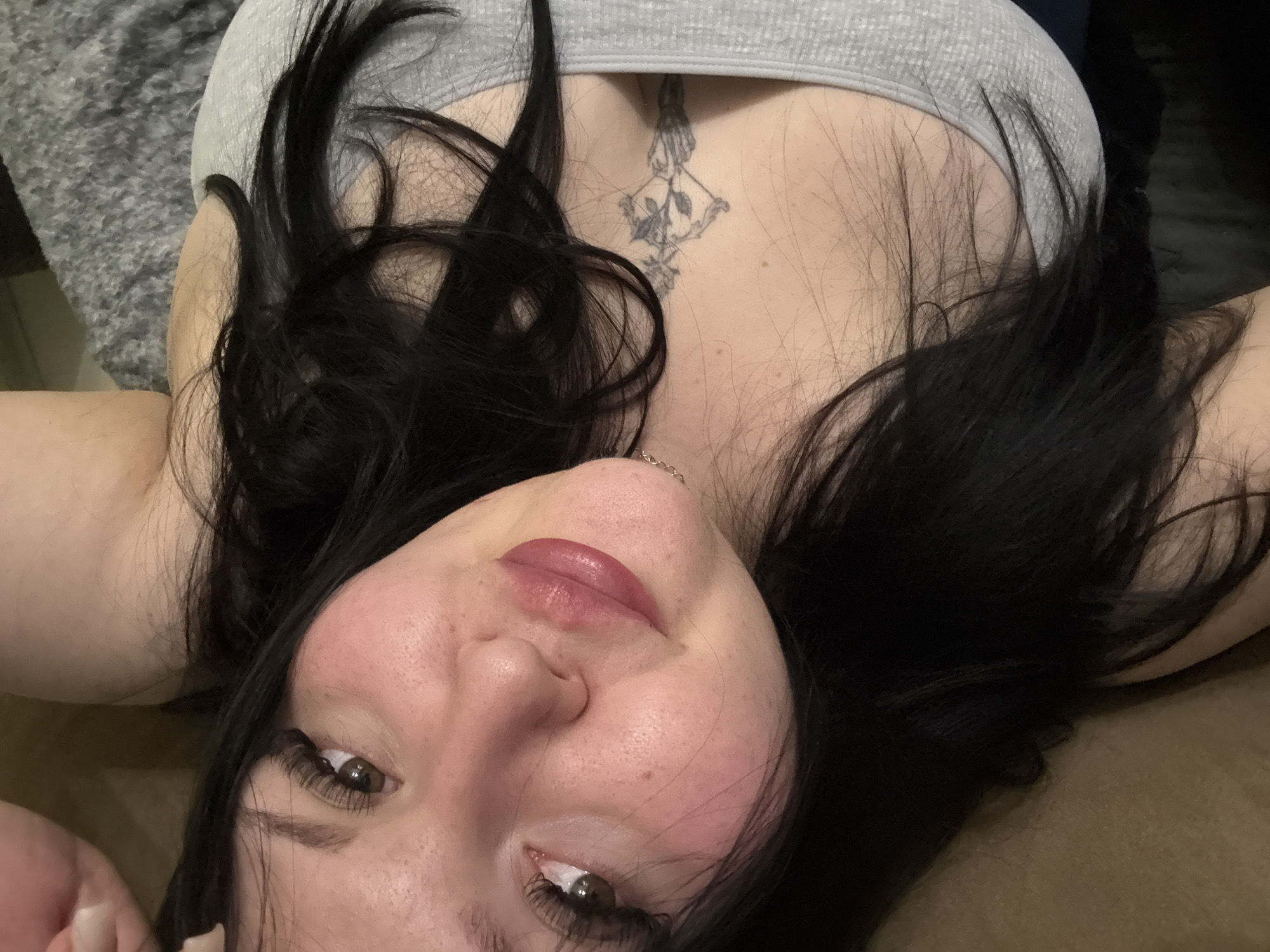 A woman with long, dark hair and a light complexion is lying down, facing upwards. She has brown eyes, long eyelashes, and slightly parted red lips. She is wearing a light grey, low-cut top that reveals a tattoo of a bow and arrow on her chest. The background is blurred and shows parts of a grey patterned surface, likely fabric. There is no information to specify this image was taken in Sofia, Bulgaria.