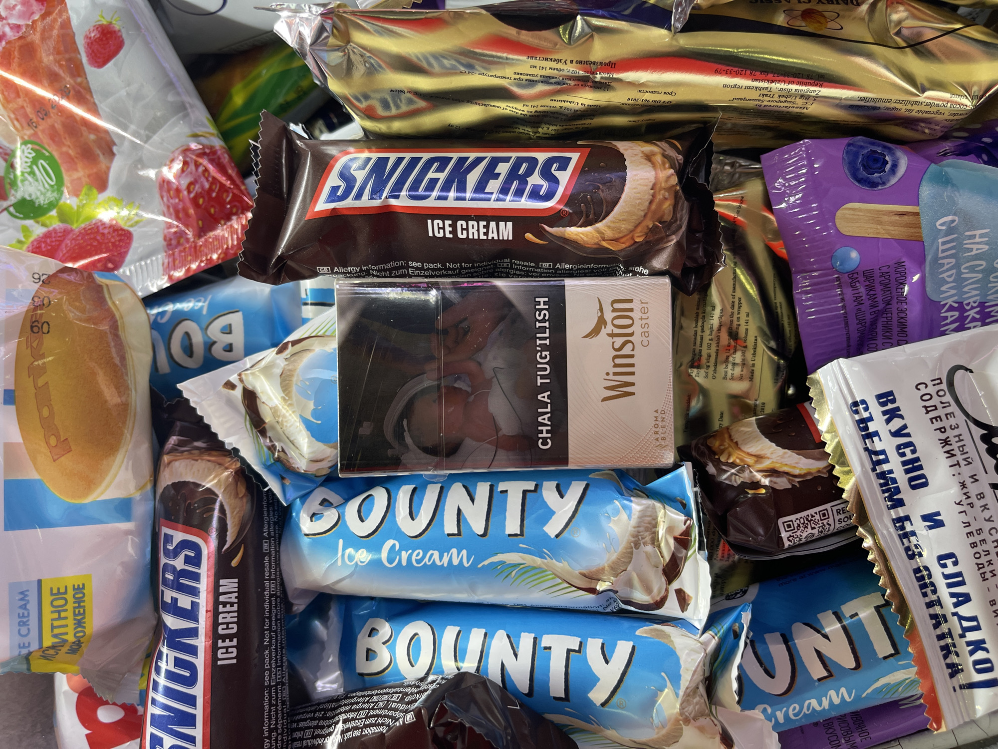 A close-up shot shows an assortment of stacked ice cream products, including Snickers, Bounty, and a blueberry-flavored popsicle. A pack of Winston caster cigarettes lies on top of the Bounty ice cream. The background includes other packaged food items. Some text is in Russian. The products appear to be from Uzbekistan, specifically manufactured by "Singapore-Samarqand" in the Zangiata district of the Tashkent region.
