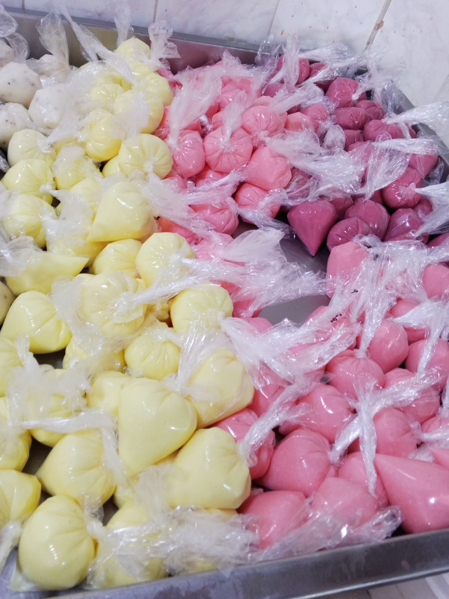 A metal tray contains small, individually wrapped plastic bags filled with various colored liquids. The liquids appear to be frozen or semi-frozen. There are four distinct color groups: white with dark specks, yellow, light pink, and dark pink/purple. The bags are tightly sealed at the top, giving them a rounded or teardrop shape. The tray is placed on what appears to be a metal countertop or shelf. The photo was taken in Tinaquillo, Venezuela.