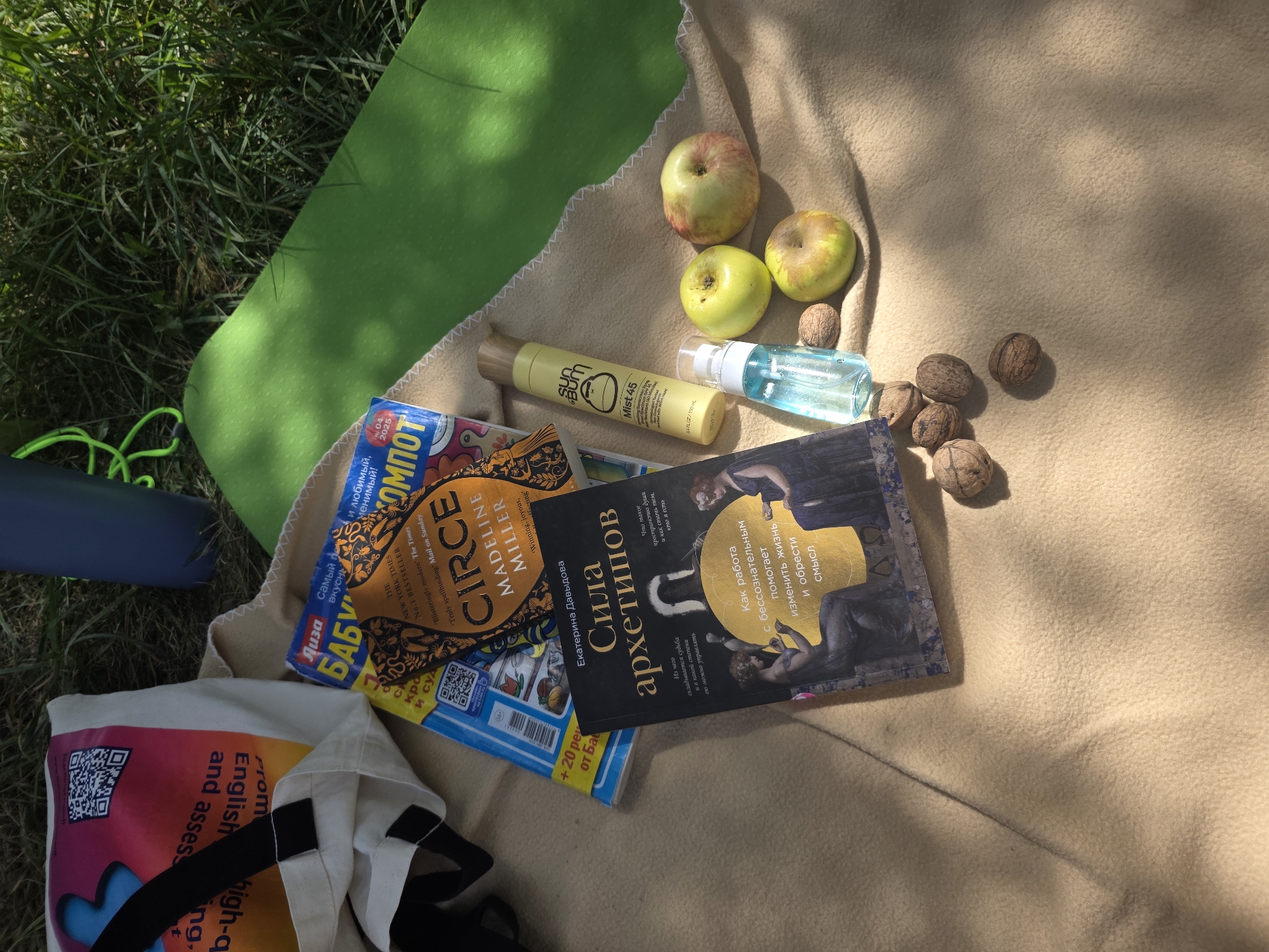 The image depicts a picnic scene laid out on a light brown blanket in a grassy outdoor environment. The objects on the blanket include two books, one titled "Сила архетипов" (The Power of Archetypes) by Екатерина Давыдова, and another book with the title "CIRCE" by Madeline Miller, along with a magazine. There are also three apples, several walnuts, a yellow spray bottle labeled "sun bum Mist 45," and a small clear spray bottle with blue liquid. In the background, a dark blue insulated bottle and a bright green yoga mat are visible, partially obscured by grass. The overall impression is that of a relaxed outdoor activity.