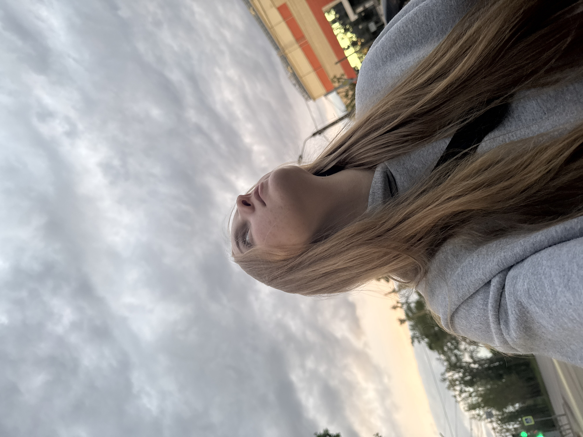 A low-angle, rotated outdoor shot depicts a young woman with light blonde, long hair, wearing a gray hooded sweatshirt. She is looking upwards towards a cloudy sky. The location appears to be an urban setting, possibly Perm, Russia, due to the visible architectural style and signage. A building with orange and white stripes is partially visible. The environment also includes trees and potentially other street furniture such as traffic lights. The image captures a moment of the woman gazing upwards in an urban setting.