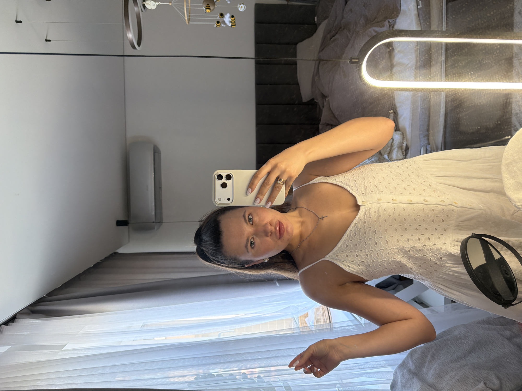 A young woman with her hair in a high ponytail, wearing a white lace sundress, takes a mirror selfie in a bedroom. She is wearing a silver necklace with a cross pendant and a ring on her left hand. The room has sheer white curtains, an air conditioning unit on the wall, and a large bed with rumpled grey bedding. A mobile with bees and clouds hangs from the ceiling, and a modern floor lamp with a glowing U-shaped light illuminates the scene. The mirror also reflects a small black makeup mirror on a stand in the foreground. The background suggests a residential interior, possibly in the city of Akulovo (Gp Kubinka), Russia.