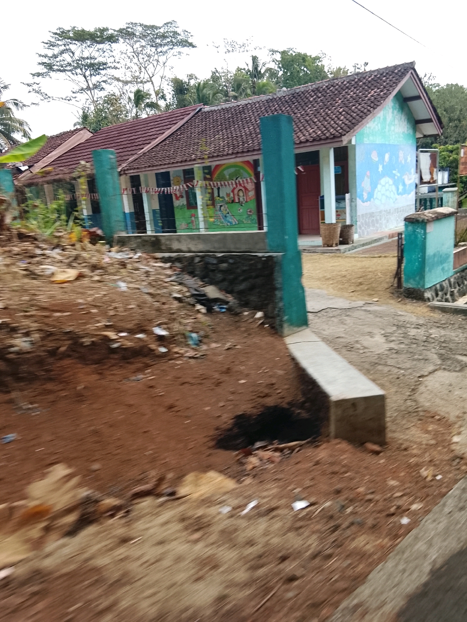 The image depicts a scene of a school building with a tiled roof, painted with bright murals of animals and space-themed imagery. The building appears to be located in a somewhat neglected or underdeveloped area, as evidenced by the presence of debris and sparse vegetation in the foreground. A concrete path leads towards the school, and there are decorative turquoise pillars visible. The overall impression is of a rural or semi-rural school in Banjar, Indonesia, possibly a kindergarten or elementary school given the colorful decorations.