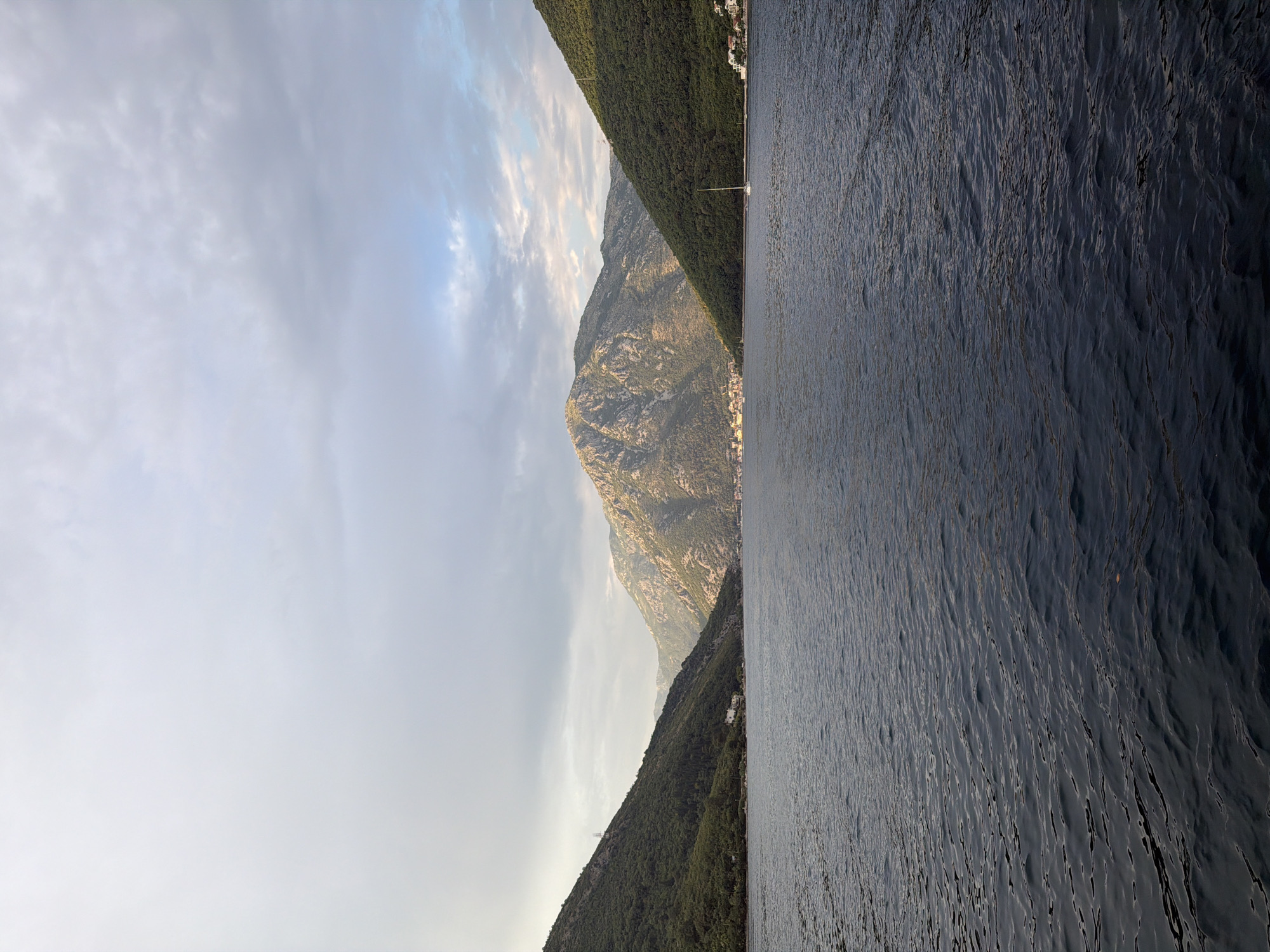 The image shows a wide shot of a body of water, likely a bay, with steep, tree-covered mountains rising on either side. A small village or town is visible nestled in the valley between the mountains in the distance. The sky is overcast with some patches of lighter sky peeking through. A single sailboat is visible on the water to the right of the frame. The overall scene suggests a serene and natural landscape. The location is identified as Lepetane, Montenegro, a settlement within the Bay of Kotor, known for its dramatic scenery and historical coastal towns.