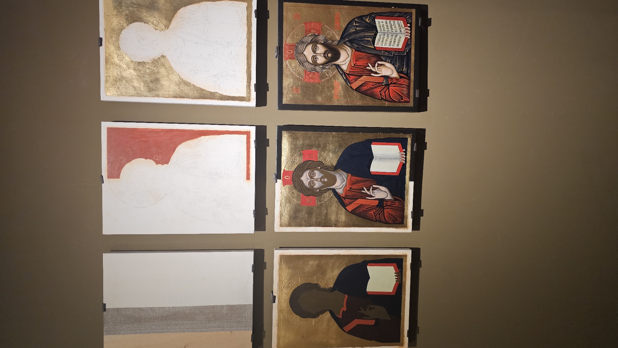 The image shows six square framed icons displayed on a beige wall. They are arranged in a 3x2 grid. The icons appear to depict a religious figure, possibly Jesus Christ, in various stages of completion.
From left to right, top to bottom:
1. An unfinished icon with a gold background and a partially outlined white figure.
2. A completed icon with a dark frame, depicting the figure holding a book.
3. An unfinished icon with a red background in the top left corner and a partially outlined white figure.
4. A partially completed icon with a gold background and the figure's face painted.
5. An unfinished icon with a white background and a layer of tan fabric at the bottom.
6. A partially completed icon with a gold background and the figure's head and body painted in dark brown.
The location of the scene is not specified, and there is no visible information to indicate it is in Varna, Bulgaria.