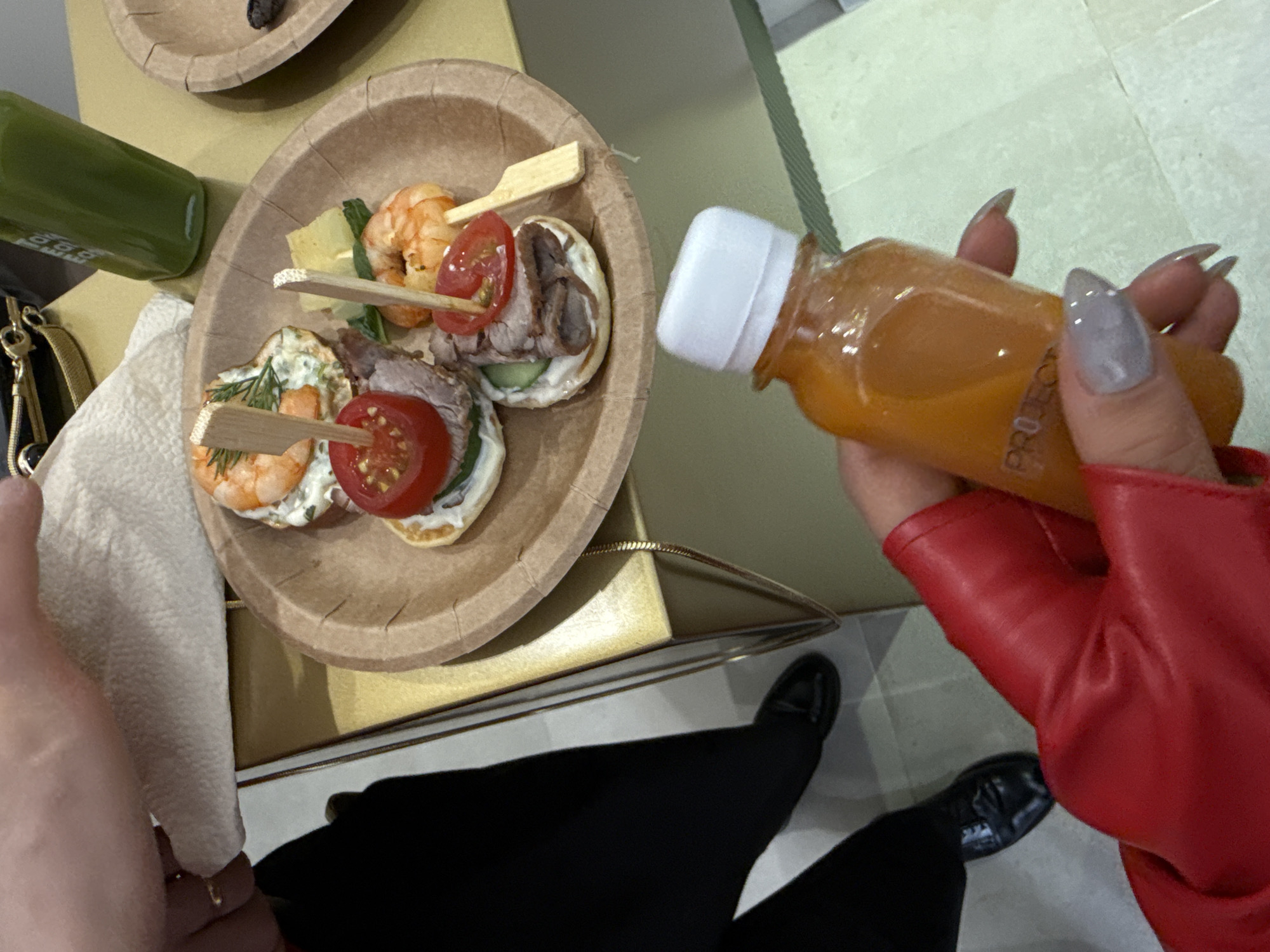 This indoor photograph, taken in Yerevan, Armenia, captures a first-person perspective at what appears to be a catered event or social gathering.
In the foreground, a person's right hand, encased in a vibrant red leather glove, holds a small, clear plastic bottle containing orange juice. The word "PROTECT" is partially visible on the bottle's label. The hand features long, pointed fingernails with a light grey polish. On the left side of the frame, a portion of the person's left hand is visible, holding a white paper napkin.
At the center of the image, resting on a light brown surface, is a round, light brown paper plate filled with various appetizers. These include small canapés, some topped with plump cooked shrimp and a sprig of dill or rosemary, while others feature thinly sliced meat (likely roast beef), cucumber, and a slice of cherry tomato. Each appetizer is secured with a small wooden skewer. Another small, green juice bottle is partially visible to the left of the plate.
A black bag with gold hardware is also visible on the table surface, nestled near the green bottle. The lower part of the image shows the person's legs, dressed in black trousers, and their feet, wearing black slip-on shoes, standing on a light-colored tiled floor. The overall setting suggests a casual yet organized event where refreshments are being served under artificial lighting.