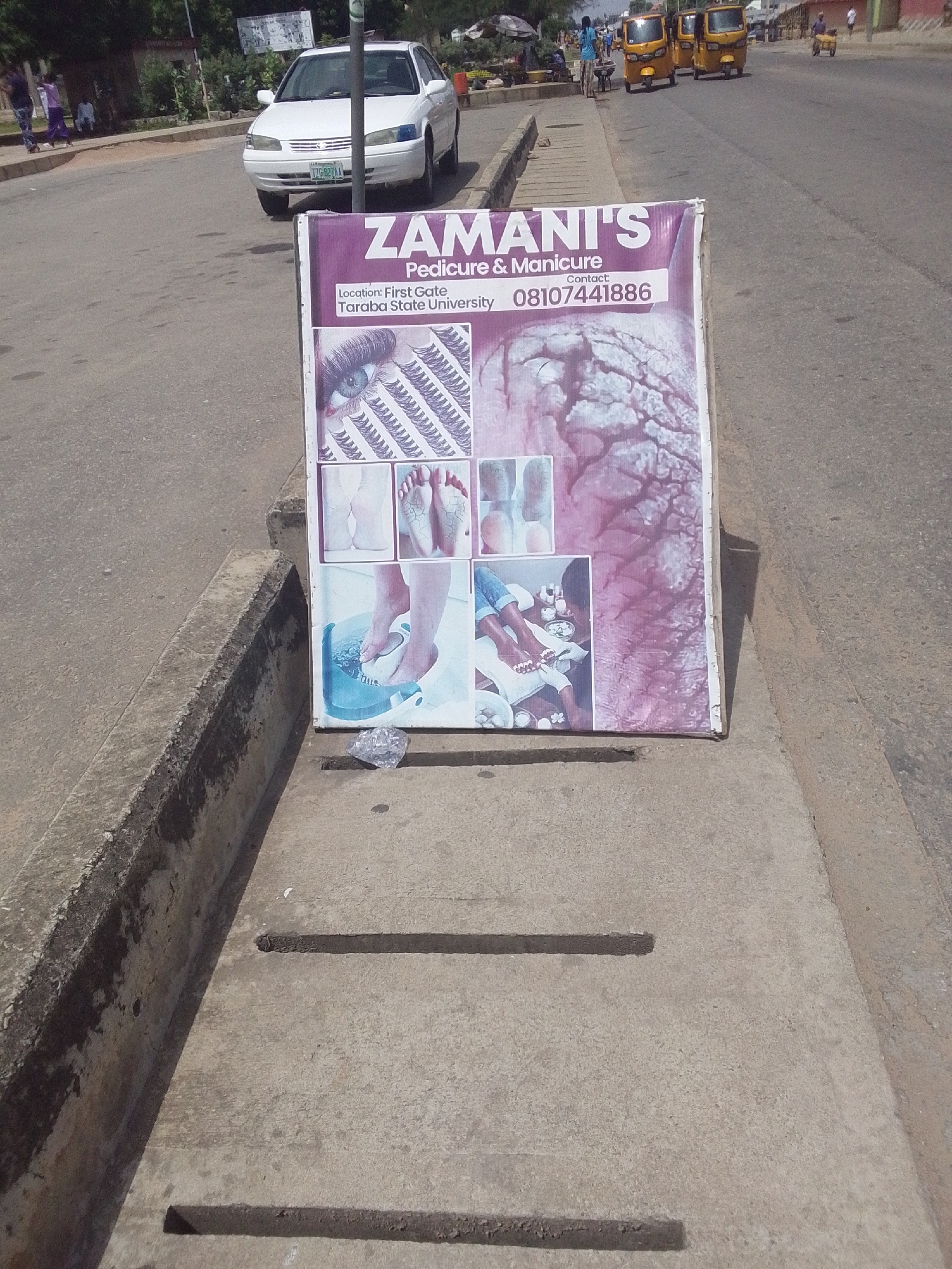 This is a medium-shot, eye-level, outdoor photograph taken on a sunny day. The scene is a street in Jalingo, Nigeria, with a white car parked on the asphalt road in the background. A purple and white freestanding sign advertising "ZAMANI'S Pedicure & Manicure" is prominently displayed in the foreground, leaning against a concrete drainage channel. The sign features images of feet, nails, and a close-up of an eye with meticulously applied eyelashes. Text on the sign indicates the location as "First Gate, Taraba State University" and provides a contact number, "08107441886". In the distance, several yellow auto-rickshaws, also known as "Keke Napep," are visible on the road, along with a few people walking. The overall impression is of a busy urban street scene, with the advertisement for a beauty service being a central focus. The lighting suggests it is daytime, and there are no strong indications of weather conditions beyond a typical sunny day.