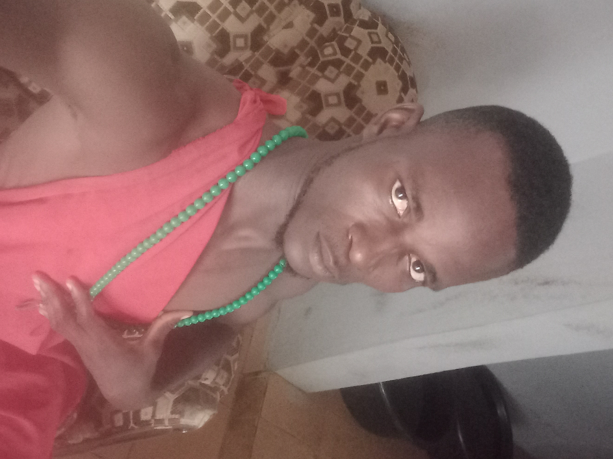 A young Black man with short hair and dark eyes is pictured from the chest up, seated in an armchair with a patterned cushion. He is wearing a bright red garment draped over his left shoulder and chest, and a strand of green beads hangs around his neck, resting on the fabric. His right arm is bent, with his hand resting on his chest. The background is a neutral-toned wall and a dark object to the right of the frame. The lighting is dim, casting shadows on the man's face and the surrounding environment.