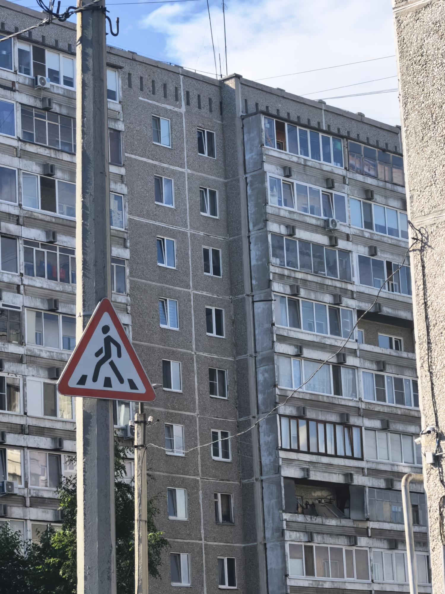 This media file shows a tall, gray, apartment building with many windows and balconies. A pedestrian crossing sign is prominently displayed on a pole in the foreground, with a silhouetted figure walking across stripes. The building appears to be made of concrete and has a utilitarian, somewhat weathered appearance. Some windows are open, and air conditioning units are visible on some balconies. The sky above is blue with some clouds, suggesting it is daytime. The overall scene suggests an urban or suburban residential area. There is no visible text in the image, aside from what might be on the signage, which is not legible. The image conveys a sense of everyday life in a developed area, with a focus on the architectural elements of the building and the traffic safety sign.