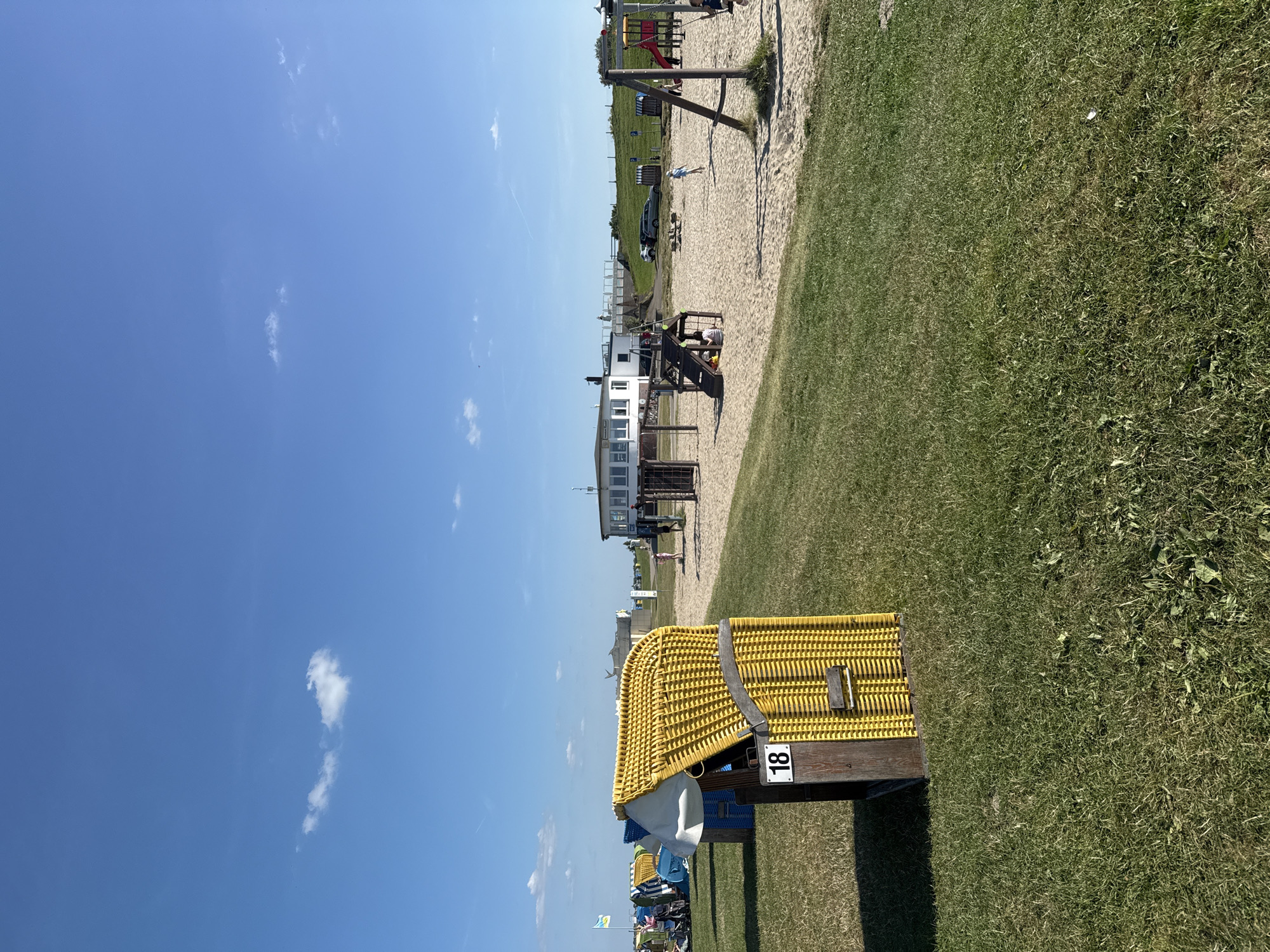 The image depicts a sunny day scene at a beach playground located in Butjadingen, Germany. In the foreground, a yellow wicker beach chair with the number "18" displayed on its side is prominently positioned on a grassy area. Beyond the chair, a sandy playground is visible, featuring a swing set with a red slide, a wooden climbing structure with stairs, and various beach chairs and tents. A building with a white facade and dark windows, likely a restaurant or visitor center, is situated in the background. Several cars are parked on a paved area behind the playground. The sky is clear blue with a few scattered white clouds. People are present, with some children playing in the playground and others relaxing in the beach chairs. The overall environment suggests a recreational area designed for families to enjoy the seaside.
