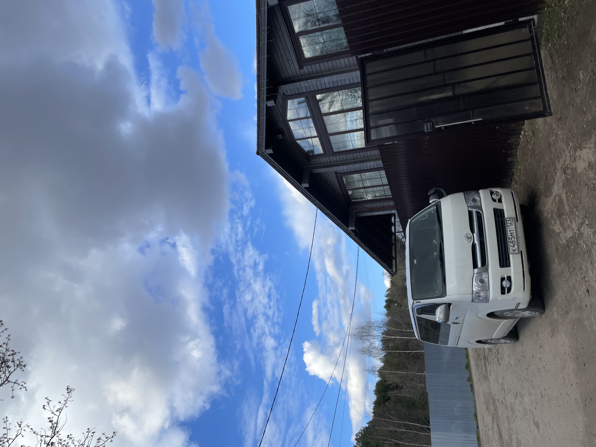 The image, presented in an upright orientation, depicts an outdoor scene featuring a white van parked on a dirt or gravel surface next to a dark brown wooden building. The building appears to be multi-story, characterized by numerous large windows, some with grid patterns, reflecting the bright, cloudy sky. Dark eaves with what look like small lights or decorations line the roof. A dark metal gate or garage door is also visible adjacent to the building.
The sky is partly cloudy, with significant areas of clear blue interspersed with white and grey cumulus clouds. In the distance, a line of deciduous trees, mostly bare, suggests the season is either early spring or late autumn. A low metal fence can be seen running along the ground near the trees. Power lines are visible stretching across the sky.
The vehicle is a white Toyota Hiace or a similar model van, positioned facing the viewer. Its front grille, headlights, and Toyota emblem are distinct. A Russian license plate, "K 485 MT 123," is affixed to the front bumper. The setting appears to be a rural or semi-rural property, consistent with a location like Izhevsk, Russia. No specific activity is taking place; the scene captures a still moment during daylight hours.