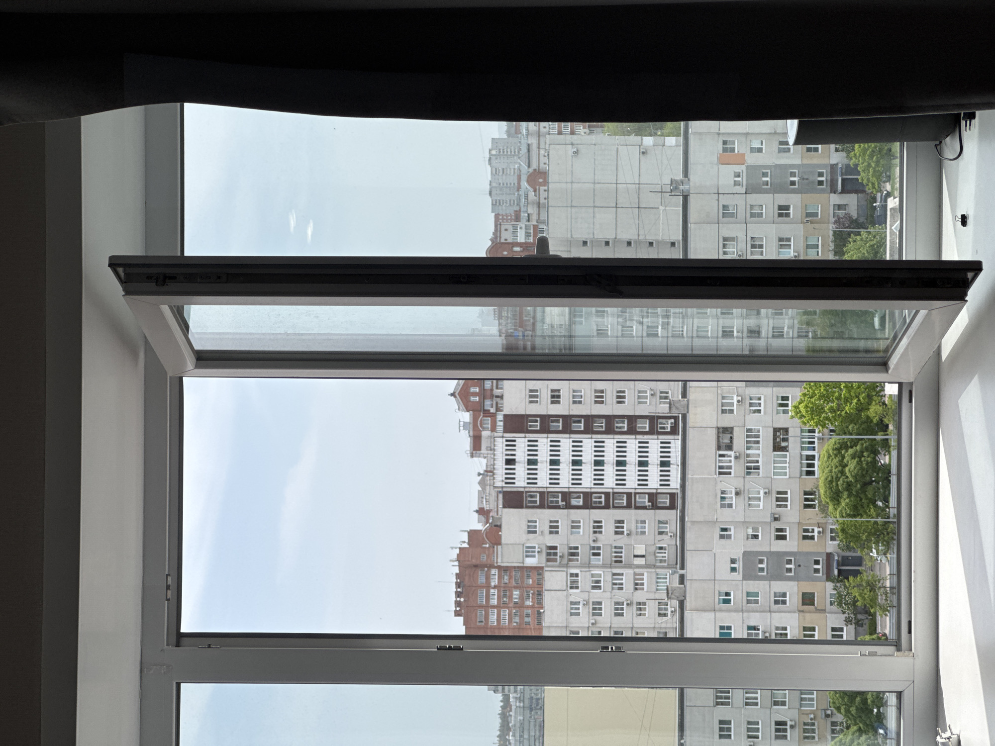 An open window frames a view of dense urban buildings under a cloudy sky. The window, white-framed with a horizontal tilt-and-turn section ajar at the top, overlooks a collection of residential buildings with repetitive window patterns and light-colored facades. Some trees are visible between the buildings. The image is taken from inside a room, with part of a dark curtain or shade visible at the top.