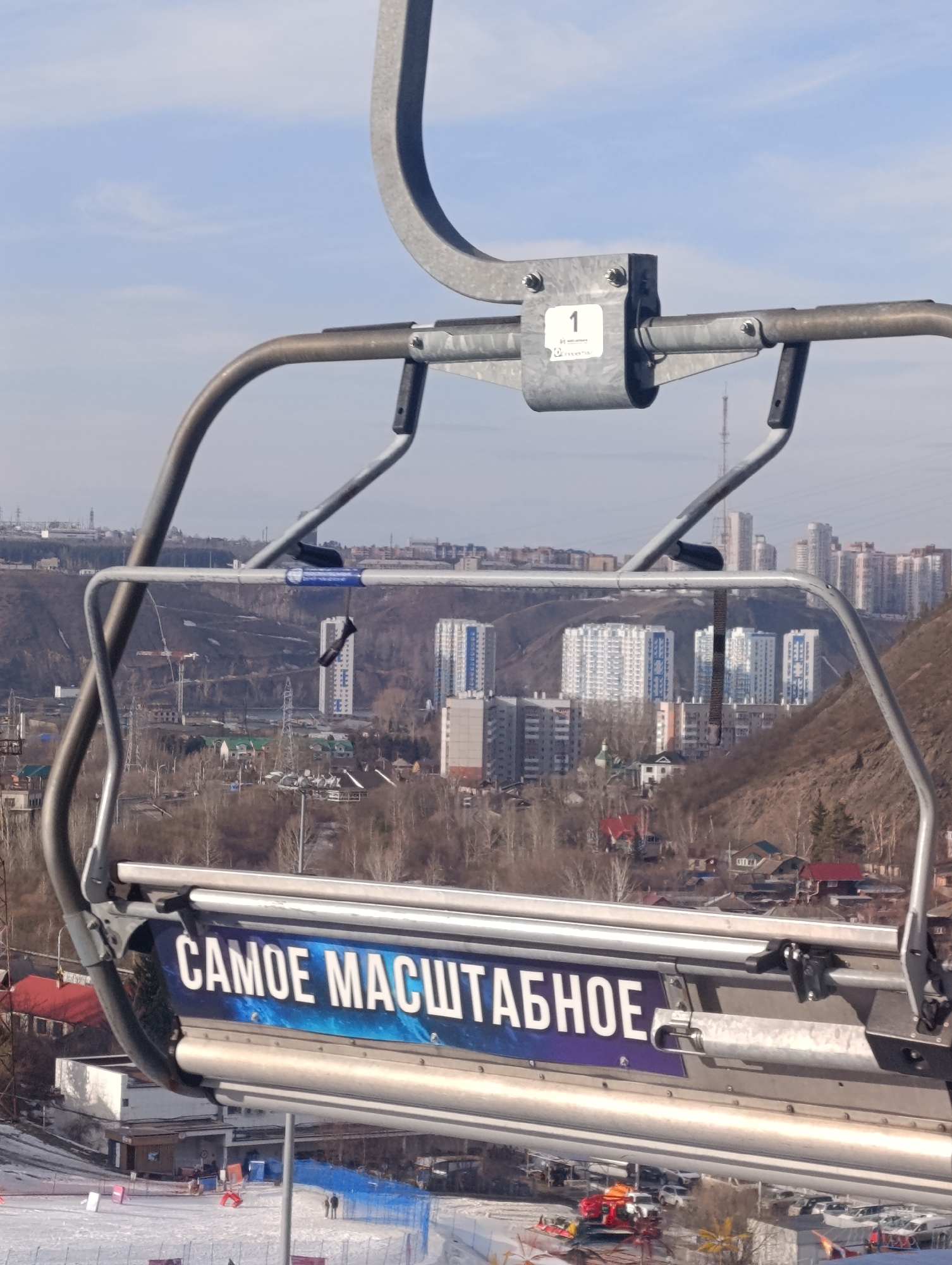 The image presents a view from a metallic ski lift chair, numbered "1", looking downhill and across a city. The chair's safety bar displays a blue and black banner with the white Russian text "САМОЕ МАСШТАБНОЕ" (THE MOST AMBITIOUS). Below the ski lift, a snow-covered ski slope is visible with blue safety netting and faint figures of individuals. A red vehicle, possibly a snow groomer or rescue sled, is on the snow, alongside a parking area filled with vehicles. The midground features a prominent hillside with bare trees and scattered residential buildings, including several modern, multi-story apartment complexes characterized by white facades and blue vertical accents. Utility poles and power lines traverse the landscape. In the background, the city of Krasnoyarsk, Russia, extends with more dense urban development and a tall communication tower visible against a pale blue sky with minimal cloud cover. The specific cluster of residential buildings and the topography indicate the scene is from the Bobrovy Log ski resort, overlooking parts of Krasnoyarsk.