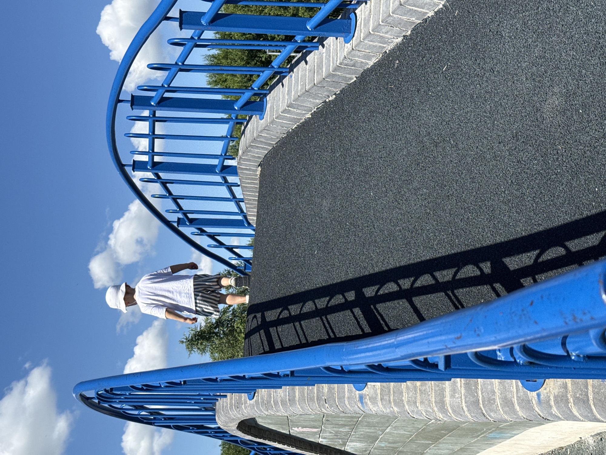 The image shows a child in a white t-shirt and striped shorts walking on a curved, dark gray surface. The child is wearing a white hat. They are walking alongside a bright blue metal railing, which curves with the surface. The railing is supported by vertical blue bars. The edge of the surface is lined with light gray bricks laid in a herringbone pattern.
The background features a bright blue sky with fluffy white clouds. Green trees are visible behind the blue railing in the distance. The sun is shining, casting a distinct shadow of the blue railing onto the dark gray surface. The shadows suggest the time of day is likely late morning or early afternoon due to the angle of the sun. The overall scene appears to be an outdoor recreational area, possibly a playground or park, given the safety surfacing and railings. The child's posture suggests they are walking casually. There is no visible text in the image.