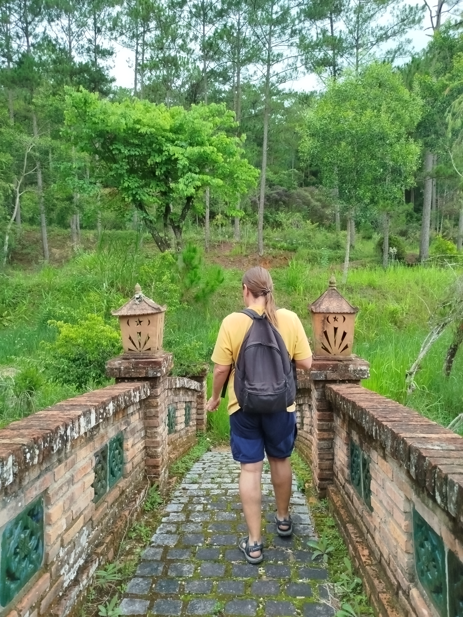 A person is seen from behind, walking across a stone and brick bridge into a verdant, forested landscape. The individual has light brown hair tied in a ponytail and is wearing a yellow short-sleeved t-shirt, blue shorts, and a dark grey backpack. Open-toed sandals are on their feet, with the brand "QUECHUA" visible on the sole of the right sandal.
The bridge features a pathway of dark grey cobblestones and walls constructed from weathered red and tan bricks, adorned with patches of moss and lichen. Green decorative ceramic elements are embedded into the brickwork along the bridge's sides. At the near end of the bridge, two light-colored, ornate lanterns with carved star and sunburst patterns rest on brick pedestals, flanking the path.
The surrounding scene is abundant with lush greenery, including tall, slender pine trees reaching skyward and various broadleaf trees with bright green foliage. Dense, vibrant green grass and smaller plants cover the ground and hillsides, creating a rich, natural environment. The lighting suggests a mild, daytime setting, possibly overcast, contributing to the soft, diffused light. The overall mood is peaceful and exploratory, set within the natural beauty of Cam Ly - Da Lat, Vietnam.