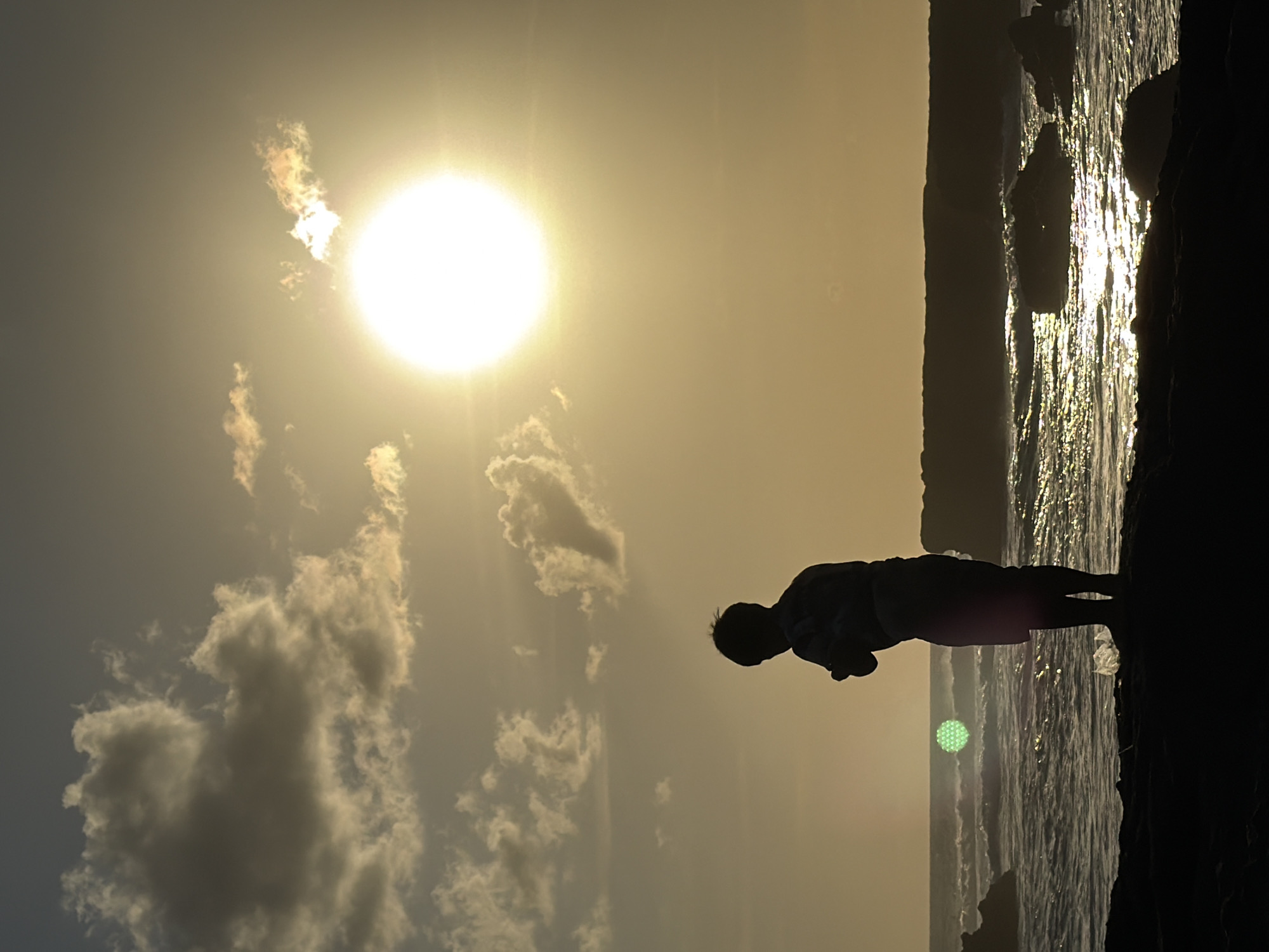 A silhouetted figure of a child stands on a dark shore, facing the ocean. The sun is setting or rising, casting a warm, golden glow across the sky and water. Clouds are scattered across the sky, some illuminated by the sun. Waves crash against the shore, and rocks are visible in the water. The child appears to be holding something in their hands. The scene is serene and evokes a sense of peace. The location is identified as North Kuta, Indonesia, suggesting a coastal setting.