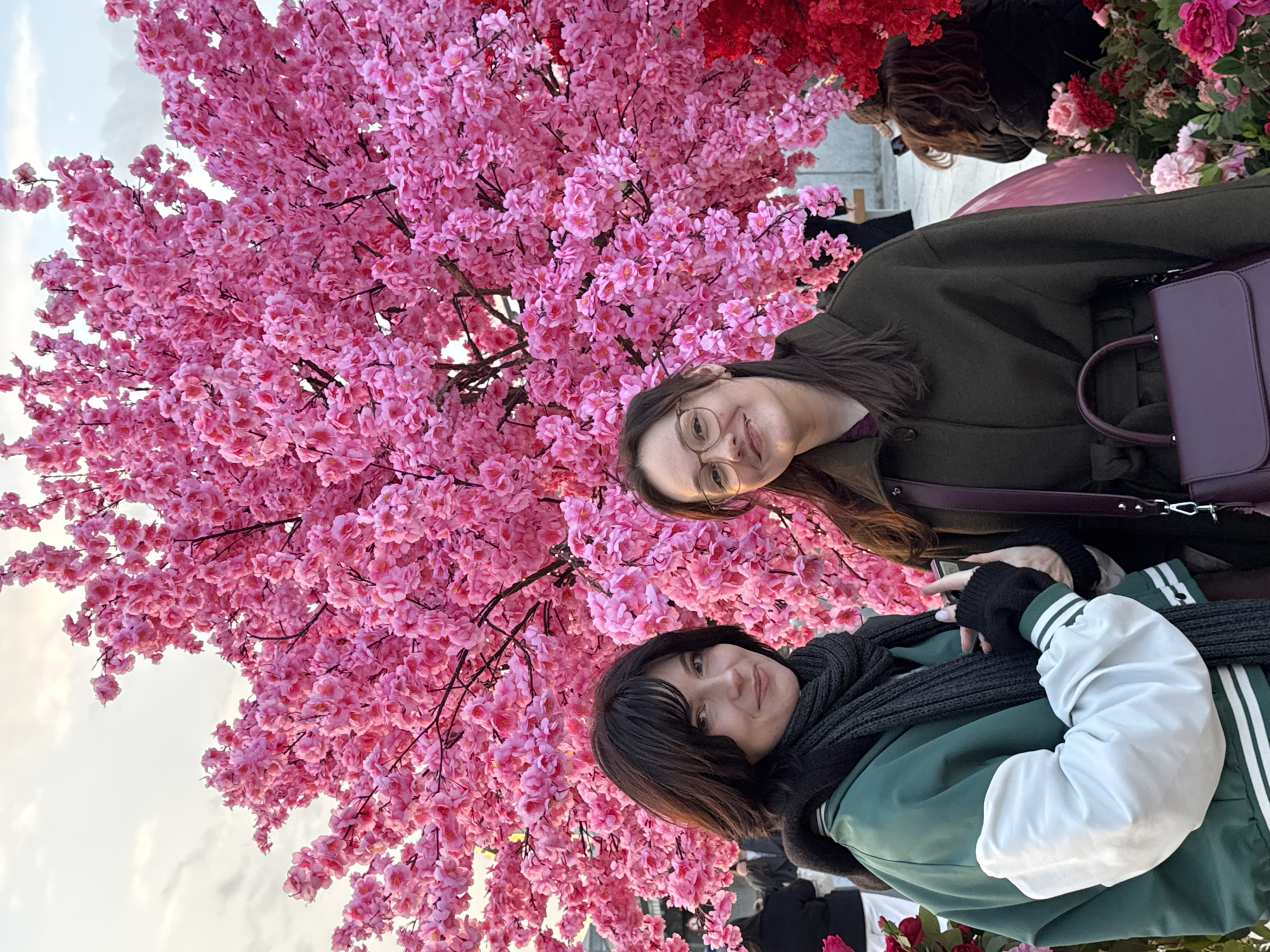Two women pose in front of a cherry blossom tree. The woman on the right wears a green jacket with white sleeves and a black scarf. The woman on the left wears glasses and a dark green coat, with a purple purse. Behind them is a dense artificial cherry blossom tree with pink flowers. Red flowers are visible in the background. The sky is overcast. The location appears to be an urban area with other people present. Based on the flowers and possible artificial construction, it could be the flower market in Moscow, Russia.