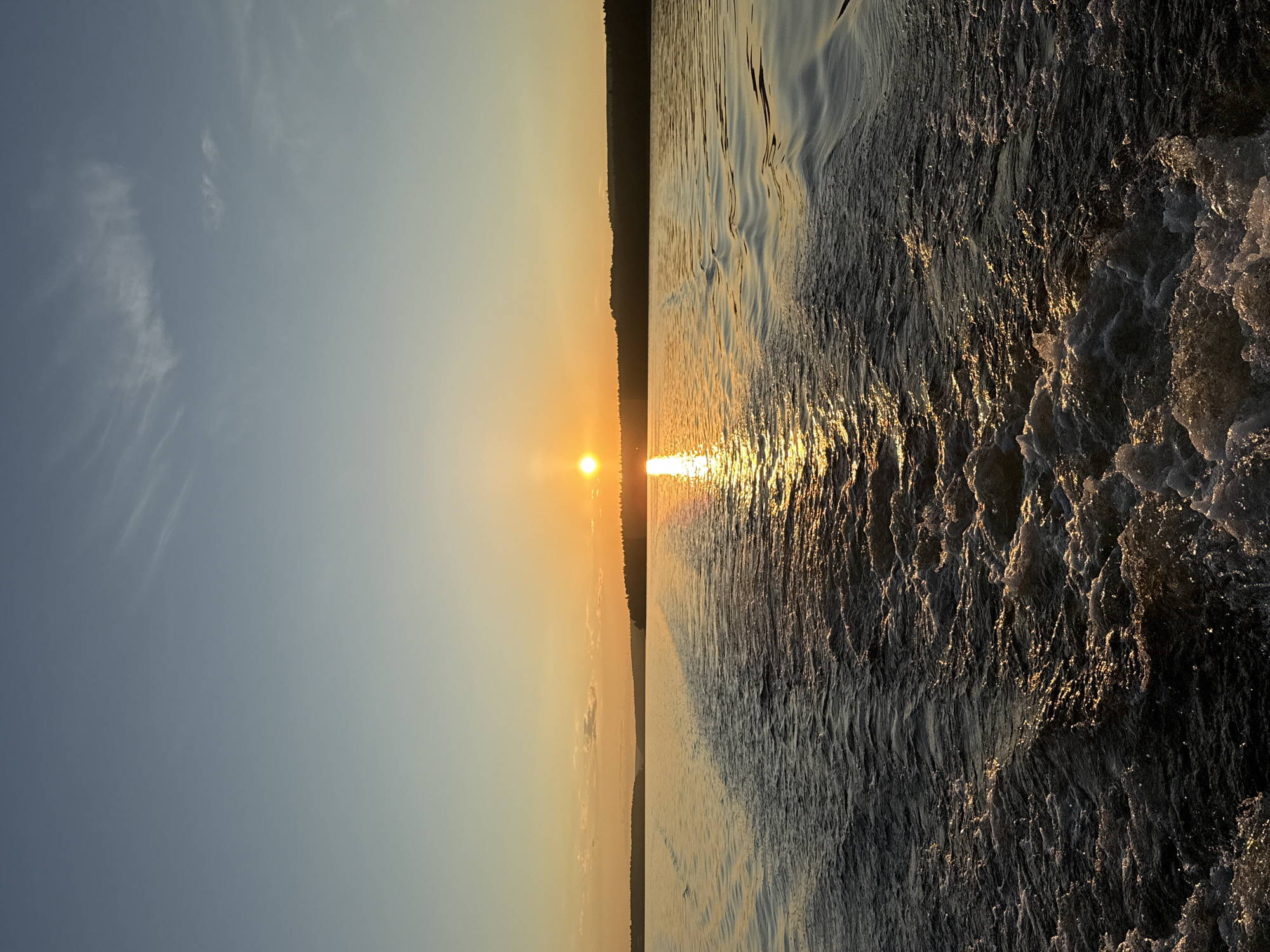 A photograph depicts a sunset or sunrise over a large body of water, identified as being located in the city of Votkinsk, Russia. The sun, a bright orange orb, is positioned low on the horizon, slightly to the right of the vertical center, casting a brilliant, shimmering golden reflection across the water directly towards the viewer. The sky above transitions from a vivid orange and yellow at the horizon to a clear, pale blue overhead, with sparse, faint cirrus clouds visible in the upper-right portion. A dark, continuous silhouette of a treeline or landmass spans the distant horizon beneath the sun.
The foreground of the image is dominated by the turbulent, frothy wake of a boat, indicating the photograph was taken from a moving vessel. This wake occupies approximately the lower half of the frame. The water within the wake is dark, almost black, but heavily churned, with white foam and spray catching the golden sunlight, creating bright, sparkling highlights against the shadowed troughs. The wake extends linearly from the bottom center of the frame towards the horizon, bisecting the sun's reflective path on the water. The calmer water to the sides of the wake exhibits subtle ripples and reflections of the sky and sun. No individuals or other distinct objects are visible within the frame apart from the natural elements and the boat's disturbed water.