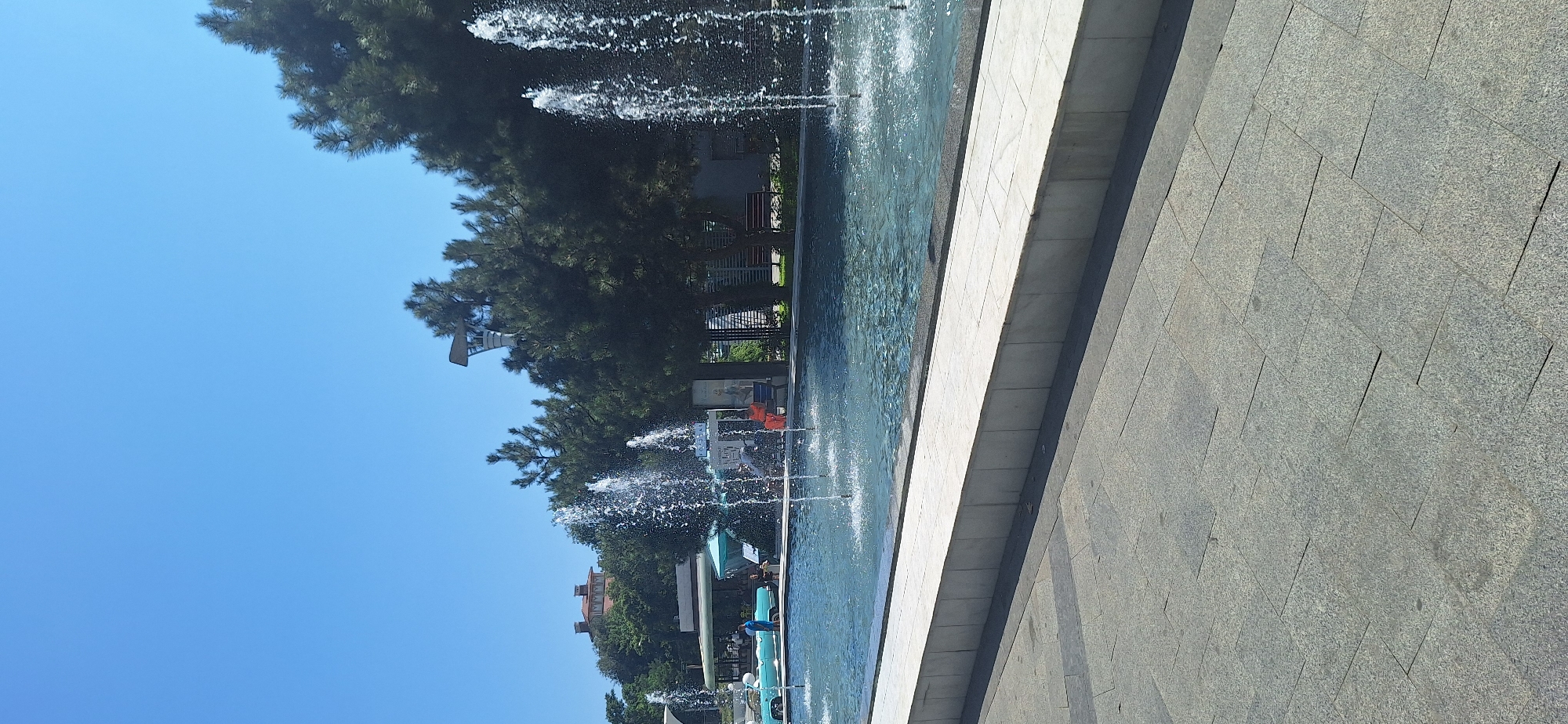 A medium shot captures an outdoor scene with a focus on a rectangular fountain filled with clear blue water. Multiple jets of water spray upwards from the fountain, creating a dynamic visual. In the background, a lush green canopy of trees forms a dense backdrop. Beyond the trees, a building with a terracotta roof is visible. The foreground features a paved walkway made of gray stone tiles. A low, white marble structure borders the fountain, separating it from the walkway. To the left of the fountain, a light blue classic car is parked. Several people are present in the scene, some standing near the car, while others are seated or standing near the fountain. The sky is a clear, bright blue, indicating a sunny day.