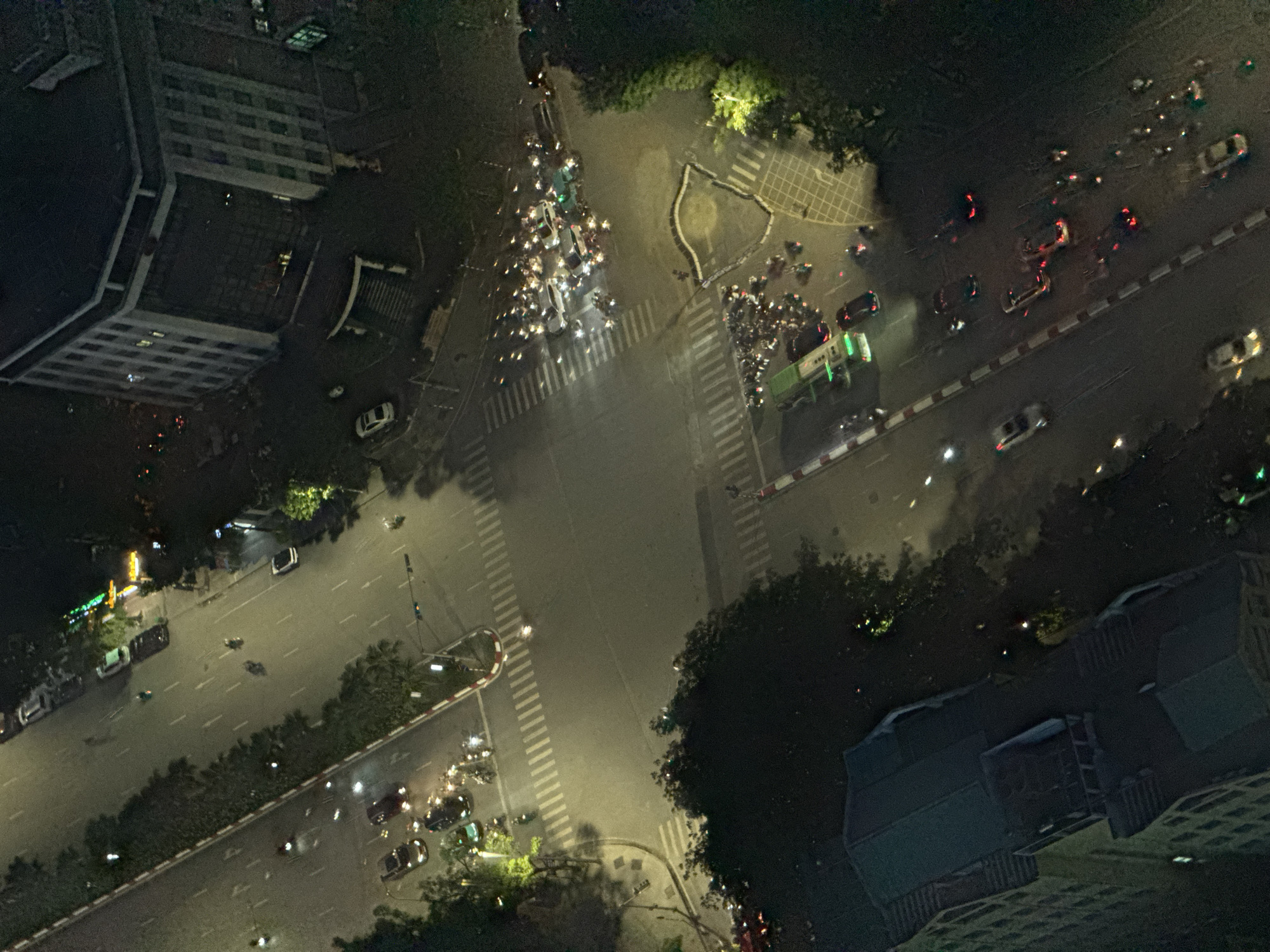 This is an aerial night shot of a busy intersection in Hanoi, Vietnam, likely near a large building with multiple floors and windows illuminated by interior lights. The intersection is characterized by heavy traffic, predominantly motorcycles, with some cars and at least one green bus. The road surface is paved, with visible white lines indicating lanes and pedestrian crossings. Streetlights and vehicle headlights cast a warm glow on the scene, creating distinct shadows. Trees line parts of the road, their dark canopies contrasting with the illuminated street. The overall atmosphere is one of vibrant urban nighttime activity.