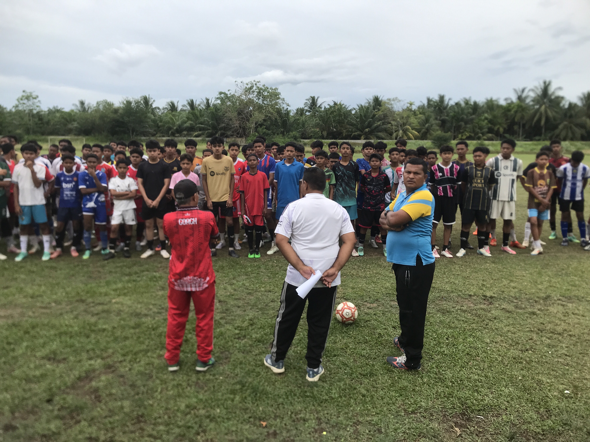 A group of male adolescents and teenagers, likely a soccer team, are gathered on a grassy field in Palohlada, Indonesia. They stand in a semi-circle facing three adult men. The adults appear to be coaches or mentors. One is wearing a red tracksuit with "COACH" printed on the back, another a white shirt with black track pants, and the third wears a blue and yellow shirt with black pants. The coach in the white shirt holds a piece of paper. A soccer ball rests on the grass near him. The environment includes a green field, trees, and a cloudy sky.
