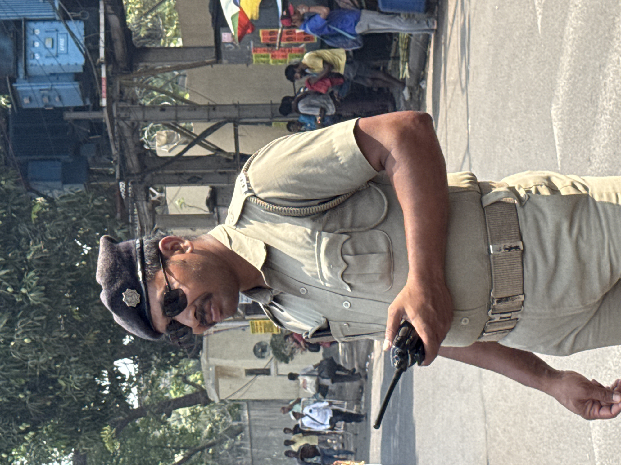 An eye-level, medium shot depicts a male police officer holding a walkie-talkie. He is wearing a khaki uniform with a black beret. The background shows a busy street in Kolkata, India, with pedestrians and various structures, including utility lines and building facades. The light suggests a sunny day.