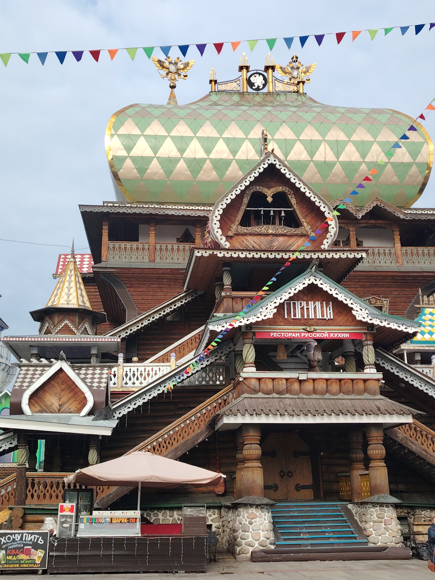 The image displays a multi-level wooden building in an elaborate traditional Russian folk architecture style, located in Moscow, Russia. The structure is richly detailed with intricate wooden carvings, decorative railings, and varied rooflines, creating a whimsical, almost fairy-tale appearance.
A prominent feature is the large, golden-domed roof, patterned with green diamond shapes. Crowning the dome is a golden double-headed eagle, a national symbol, along with other embellishments like a clock face and smaller spires. Colorful triangular bunting is strung across the sky and between different sections of the building, adding a festive touch.
The building houses a restaurant, indicated by a sign reading "ГОРНИЦА" (GORNITSA) and "РЕСТОРАН РУССКОЙ КУХНИ" (RESTORAN RUSSKOY KUKHNI), which translates to "Russian Cuisine Restaurant." Below this, a grand entrance is visible, featuring sturdy wooden pillars and stone foundations, with blue-painted wooden steps leading up to it.
At ground level, an outdoor vending setup is present. A stand beneath a beige umbrella advertises "МОРОЖЕНО ШАРИК-250" (MOROZHENO SHARIK-250), meaning "Ice cream scoop - 250" (presumably rubles). Adjacent to it, a chalkboard sign titled "ХОЧУ ПИТЬ" (KHOCHU PIT' - "I Want to Drink") lists "ПРОХЛАДИТЕЛЬНЫЕ НАПИТКИ" (PROKHLADITEL'NYE NAPITKI - "Refreshing Drinks"), including "КВАС" (Kvass), "МЕДОВУХА" (Medovukha), "ТАРХУН" (Tarragon), "МОХИТО" (Mojito), and "ДОМАШНИЕ ЛИМОНАДЫ" (Homemade Lemonades).
The sky is overcast and grey, suggesting daytime on a cloudy day. No people are prominently visible, but the commercial establishments imply an active area for dining and refreshments. The overall scene exudes cultural charm and hospitality.